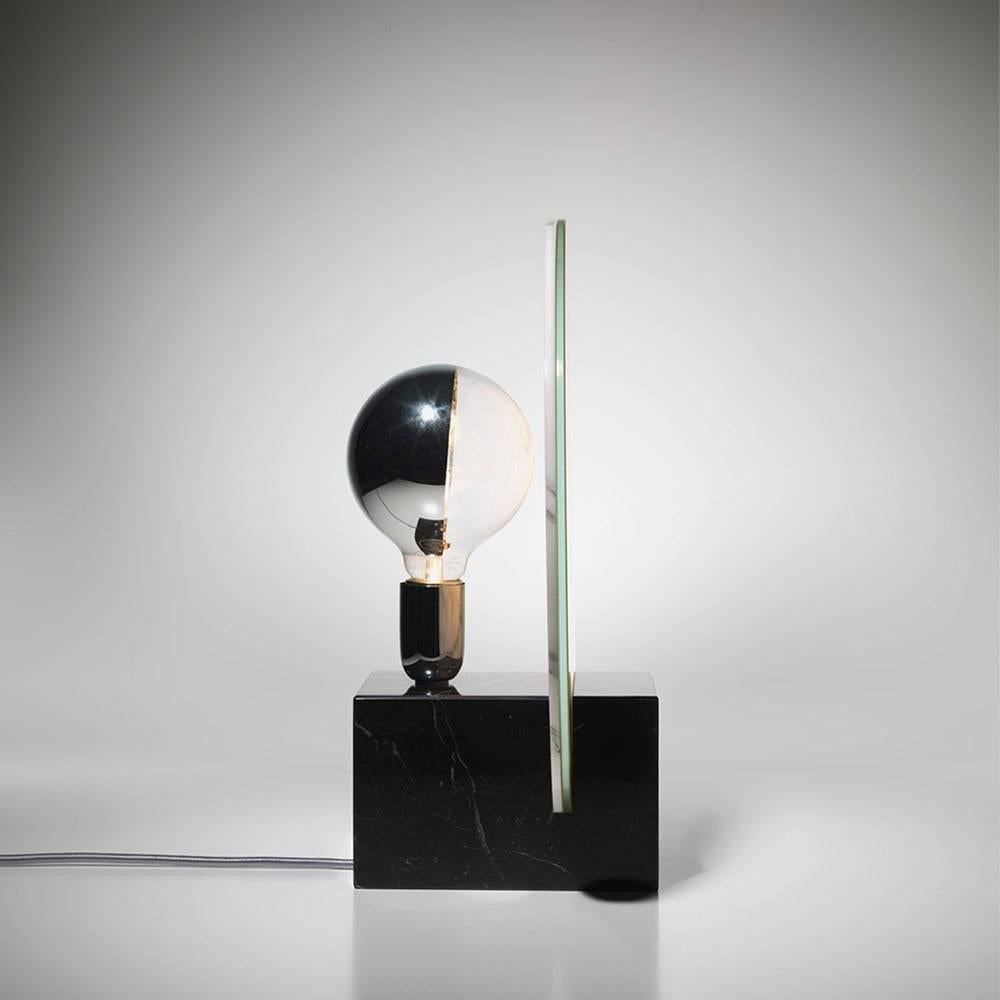 A layer of white Carrara marble and acid yellow tinted glass are sandwiched together to form an illuminated circular diffuser for this sculptural table lamp. Set into a solid black nero marquina marble base, the veins of the Carrara marble are