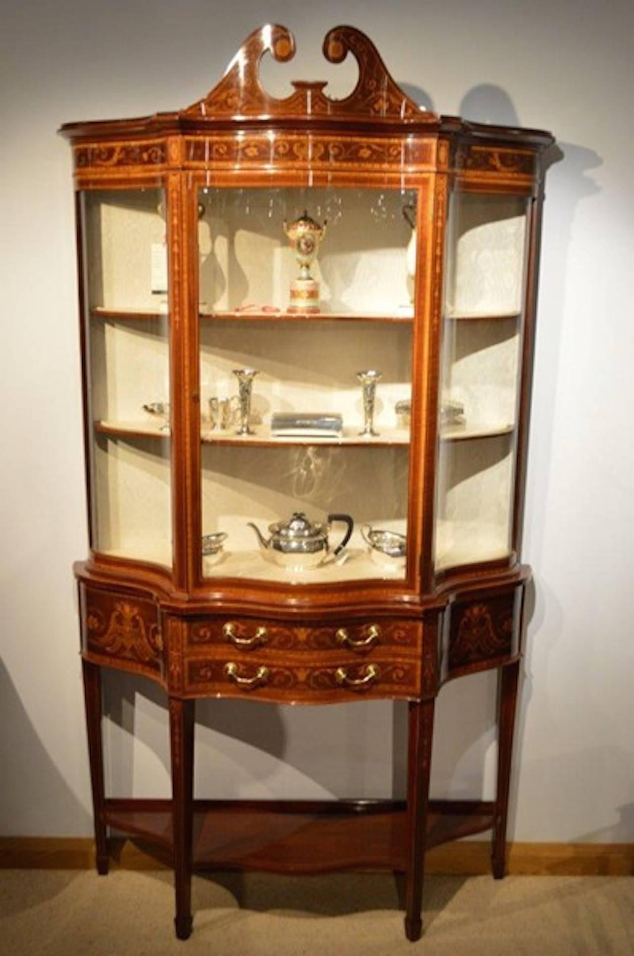 An exhibition quality mahogany inlaid serpentine antique display cabinet by Maple & Co of London. The upper section with a broken swan neck pediment with marquetry inlaid detail and circular paterae above the serpentine frieze depicting mermaids and