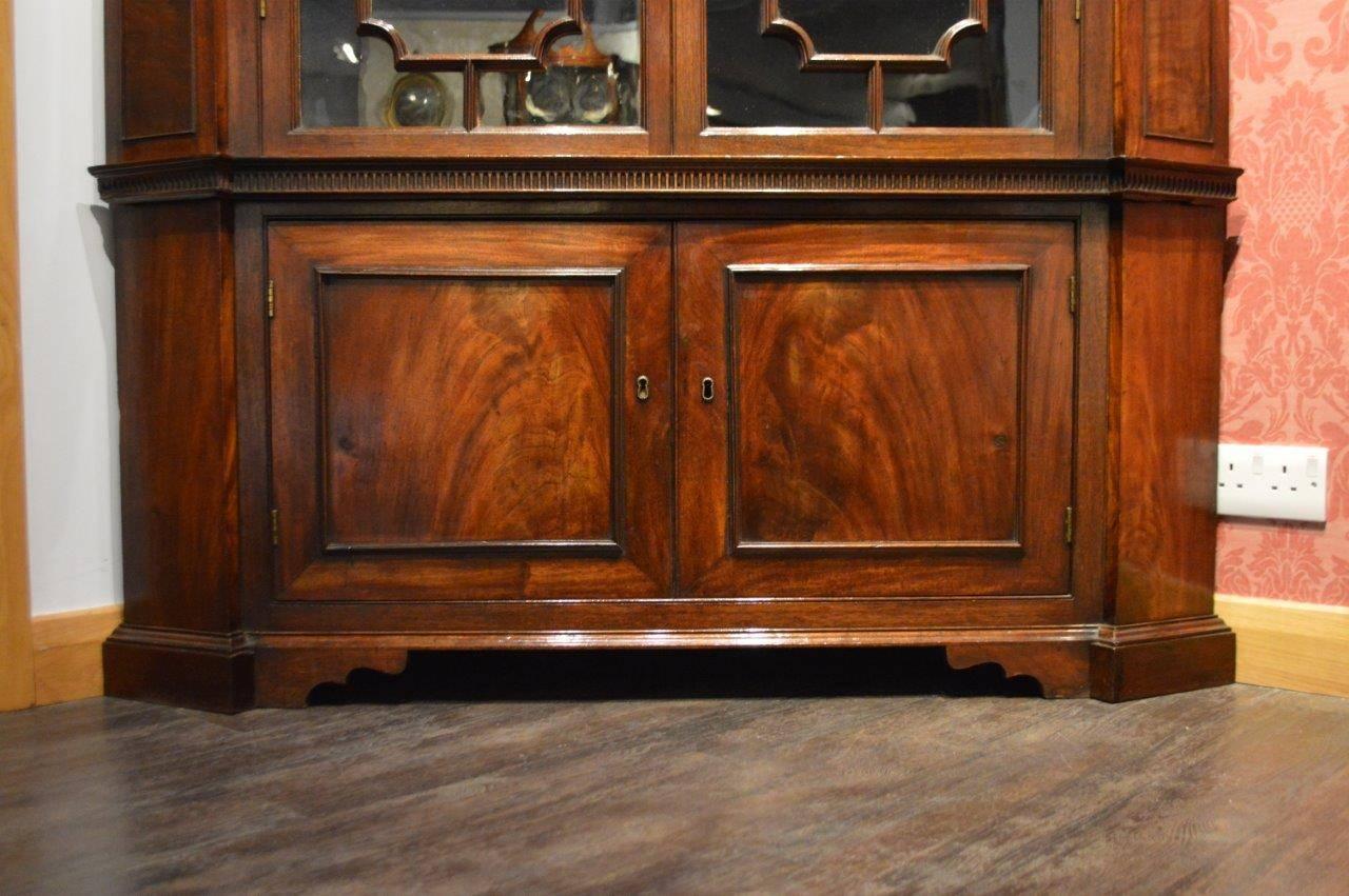 A mahogany George III Period Chippendale inspired double corner cabinet. The upper section with an inverted breakfront dentil moulded cornice above a flame mahogany veneered frieze with central carved and anthemion detail, above twin astragal glazed