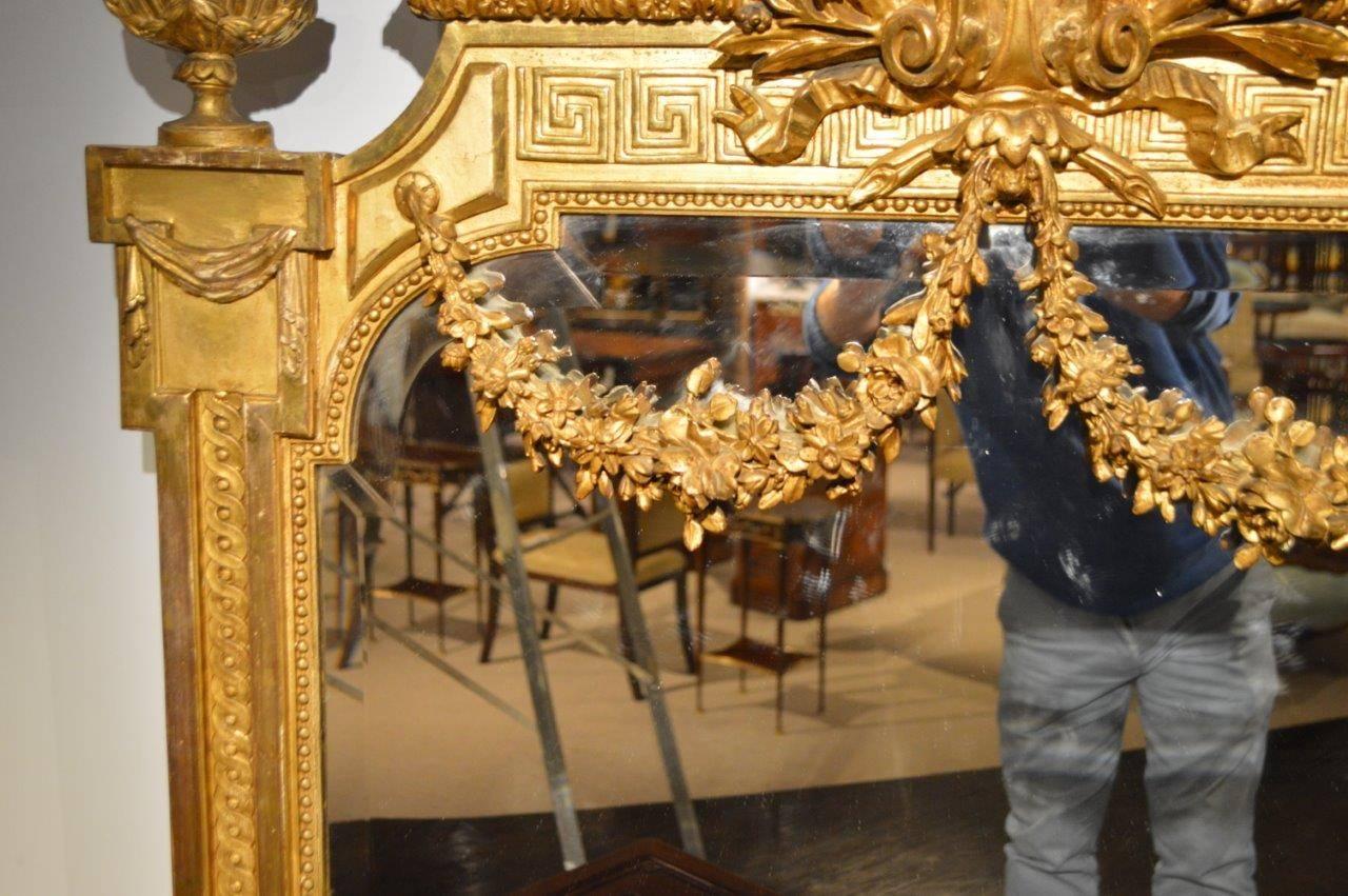 A fine quality carved giltwood late 19th century French mirror. Having an elaborately carved central cartouche flanked by classical giltwood urns and with a Greek key frieze supporting finely carved and gilded garlands. The shaped bevelled mirror