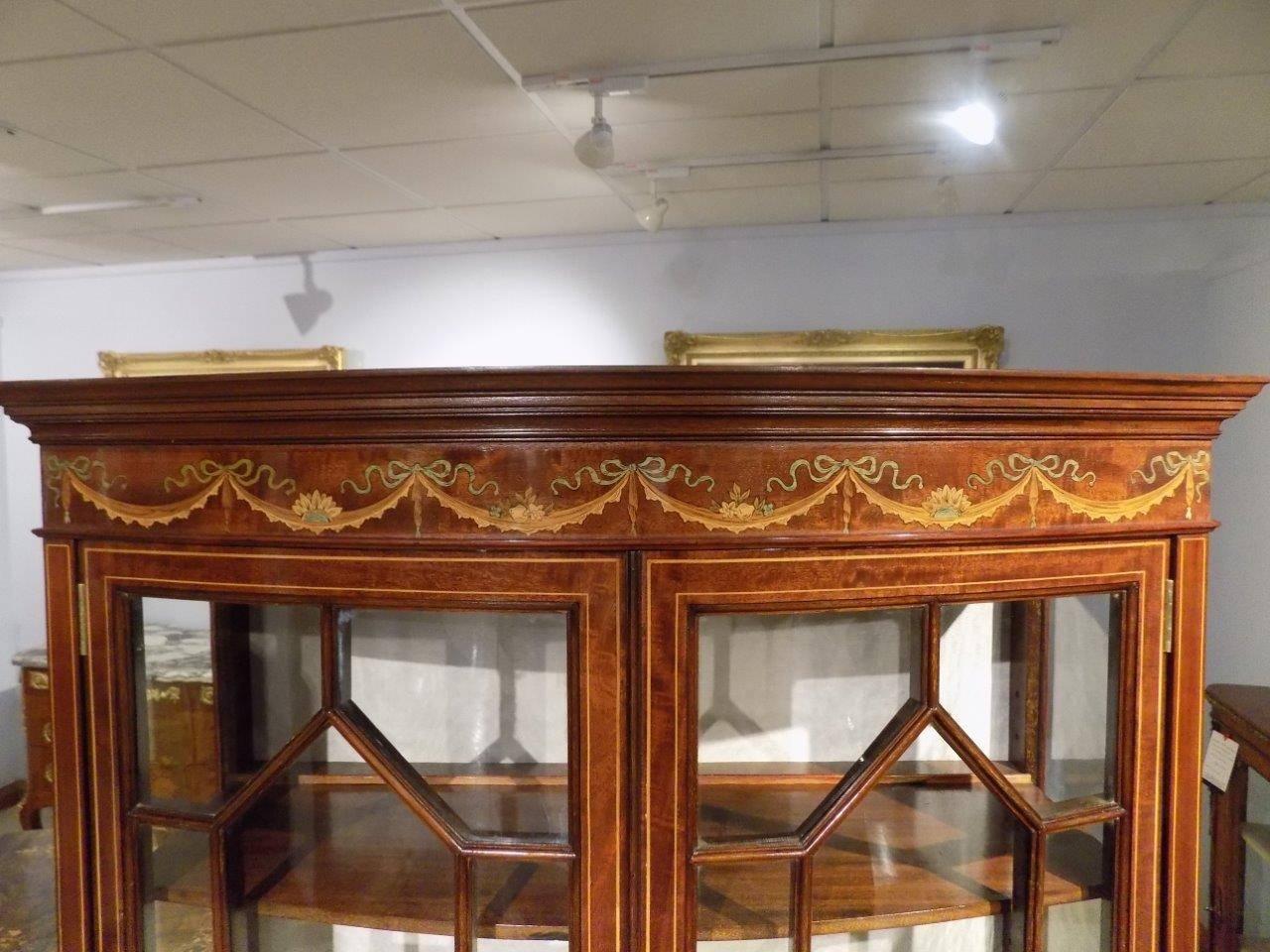 A superb quality mahogany and marquetry inlaid bow front display cabinet on stand. The upper section with a moulded cornice above a ribbon and swag marquetry and pen-work inlaid frieze with twin bow front astragal glazed doors, opens to reveal a
