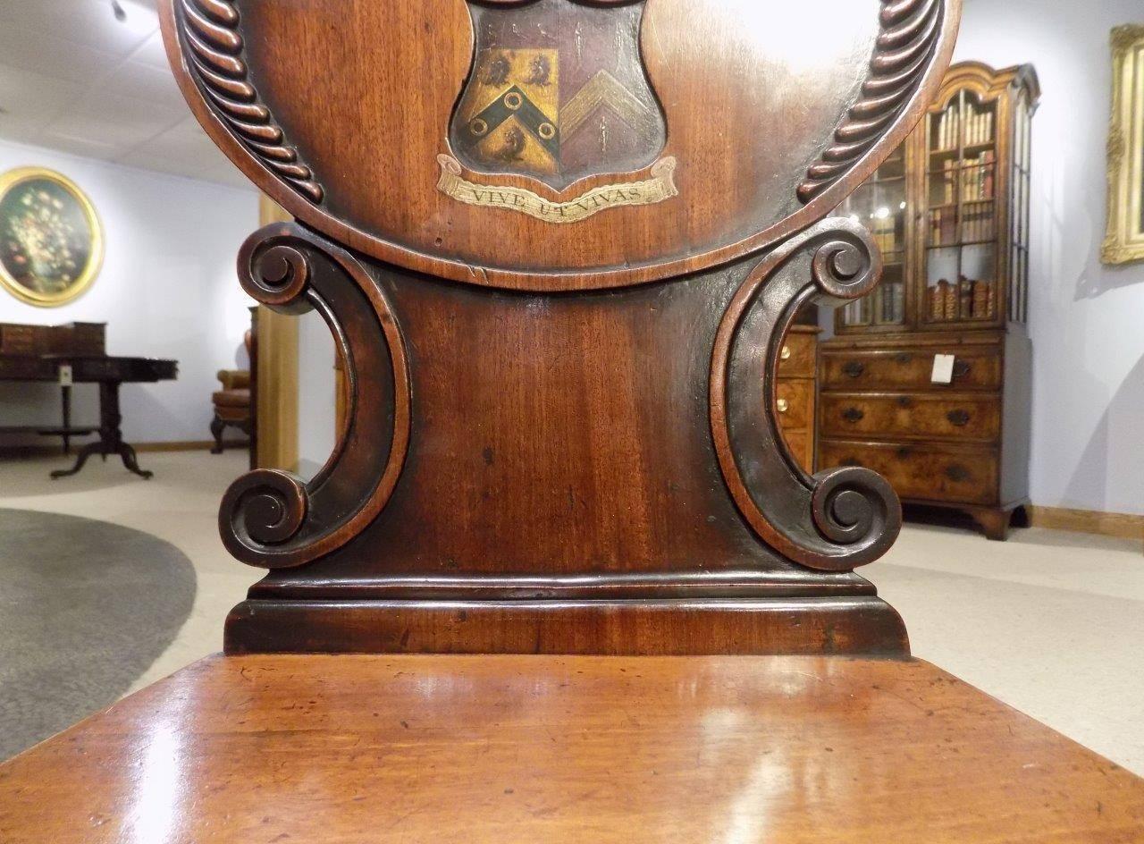 A good pair of mahogany William IV period antique hall chairs. Each with a finely carved back with a central Coat of Arms and solid mahogany seat. Supported on octagonal faceted front legs and swept rear supports, English, circa