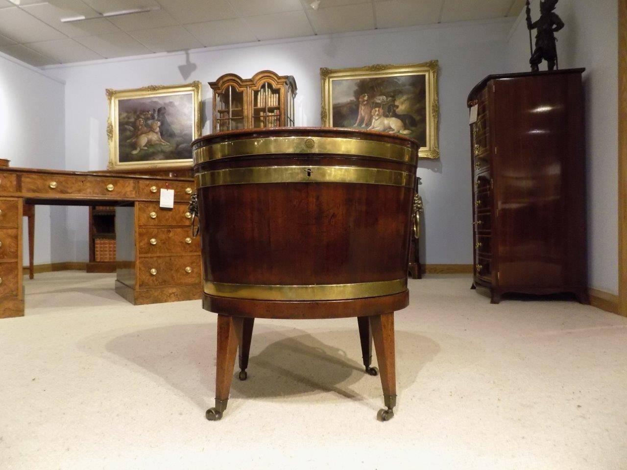 A George III period Cuban mahogany oval lead lined wine cooler. Having an oval hinged top opening to reveal a partially lead lined interior with plug. Retaining the original lions mask handles and supported on the original oval stand with square