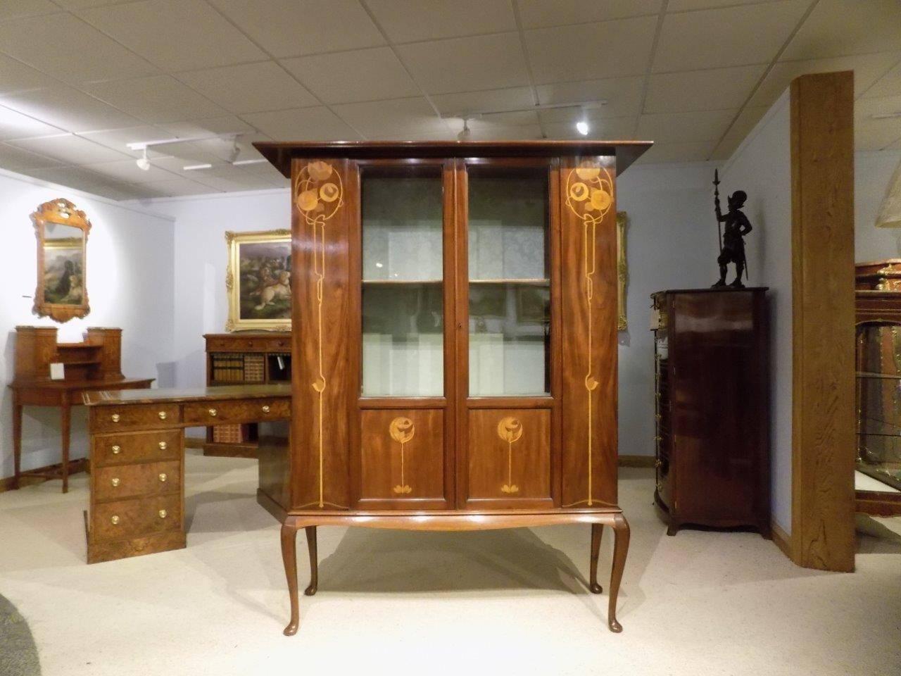 A mahogany inlaid Arts & Crafts period display cabinet by The Bath Cabinet Makers Ltd. The overhanging mahogany top above twin glazed doors with a silk lined interior with stylised Art Nouveau inlaid floral panels in the style of George Walton.