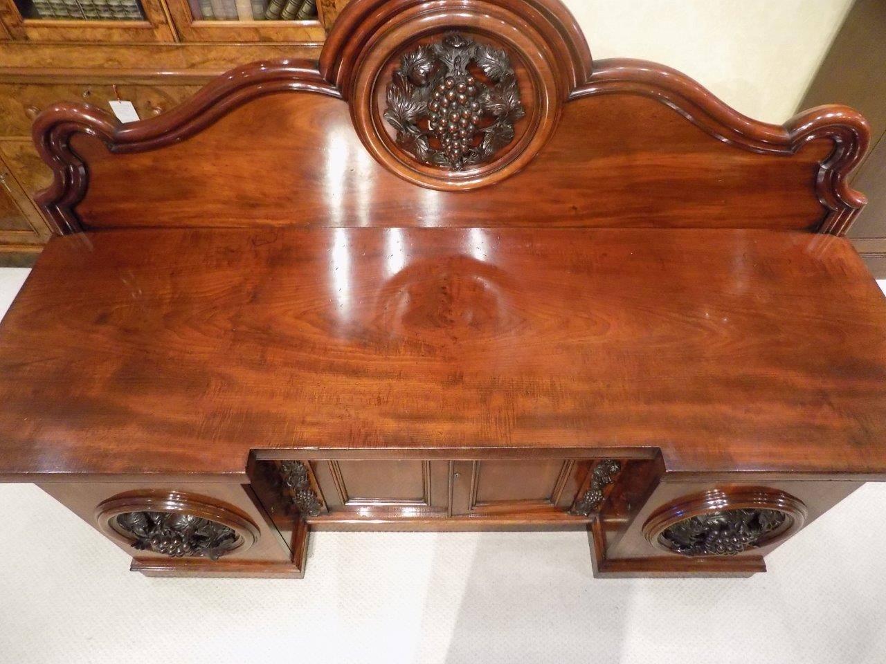 A superb quality figured mahogany Victorian period four-door antique sideboard. Having a raised arched back with a stunning carved central roundel depicting grape and vine leaves which is repeated on the doors of the base. The rectangular top of
