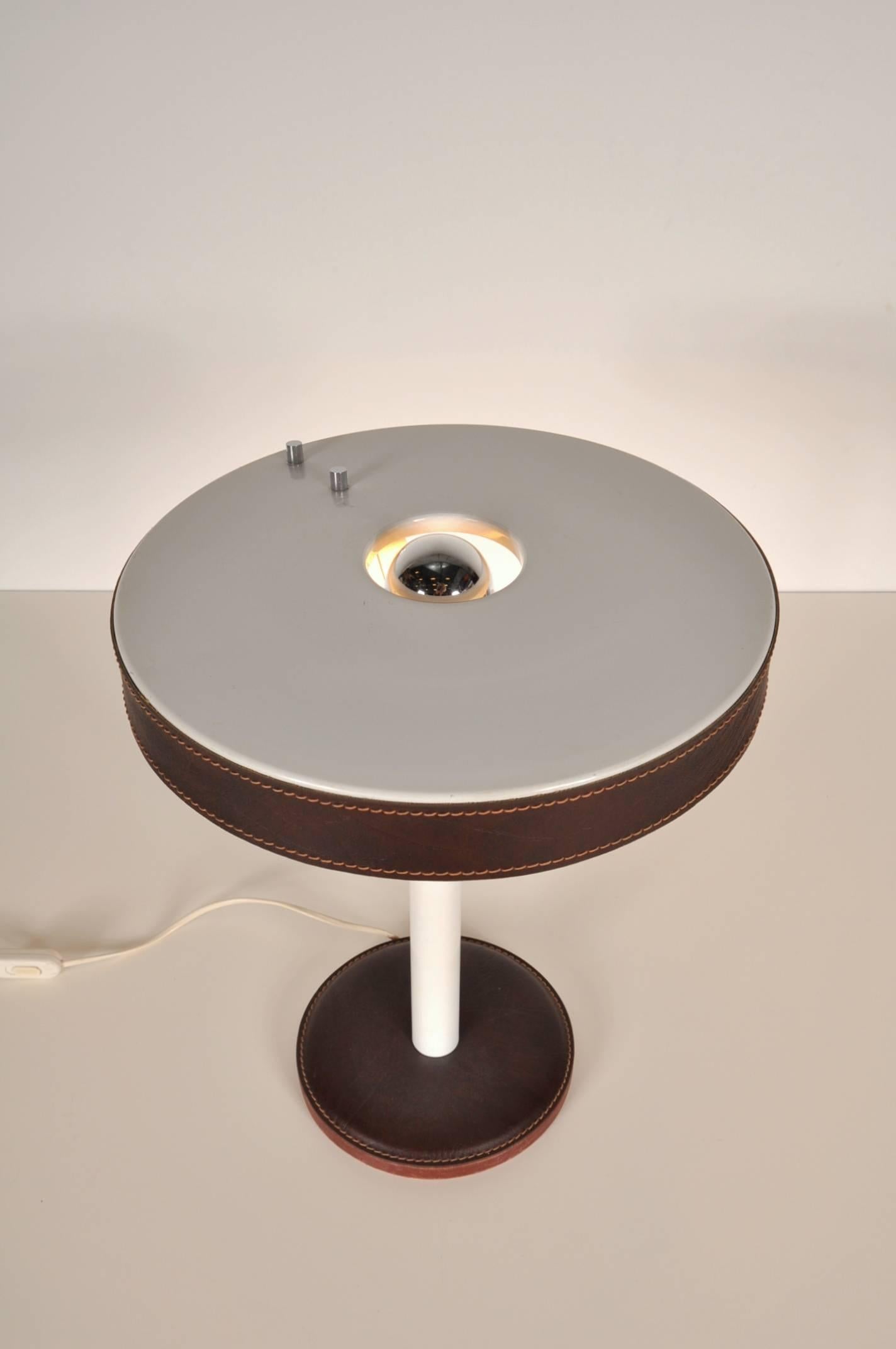Beautiful table or desk lamp designed by Jacques Adnet in France, circa 1950.

The lamp is made of very well crafted stitched brown leather, making it a highly recognizable Adnet piece. The base is made of white metal, giving it an eye-catching