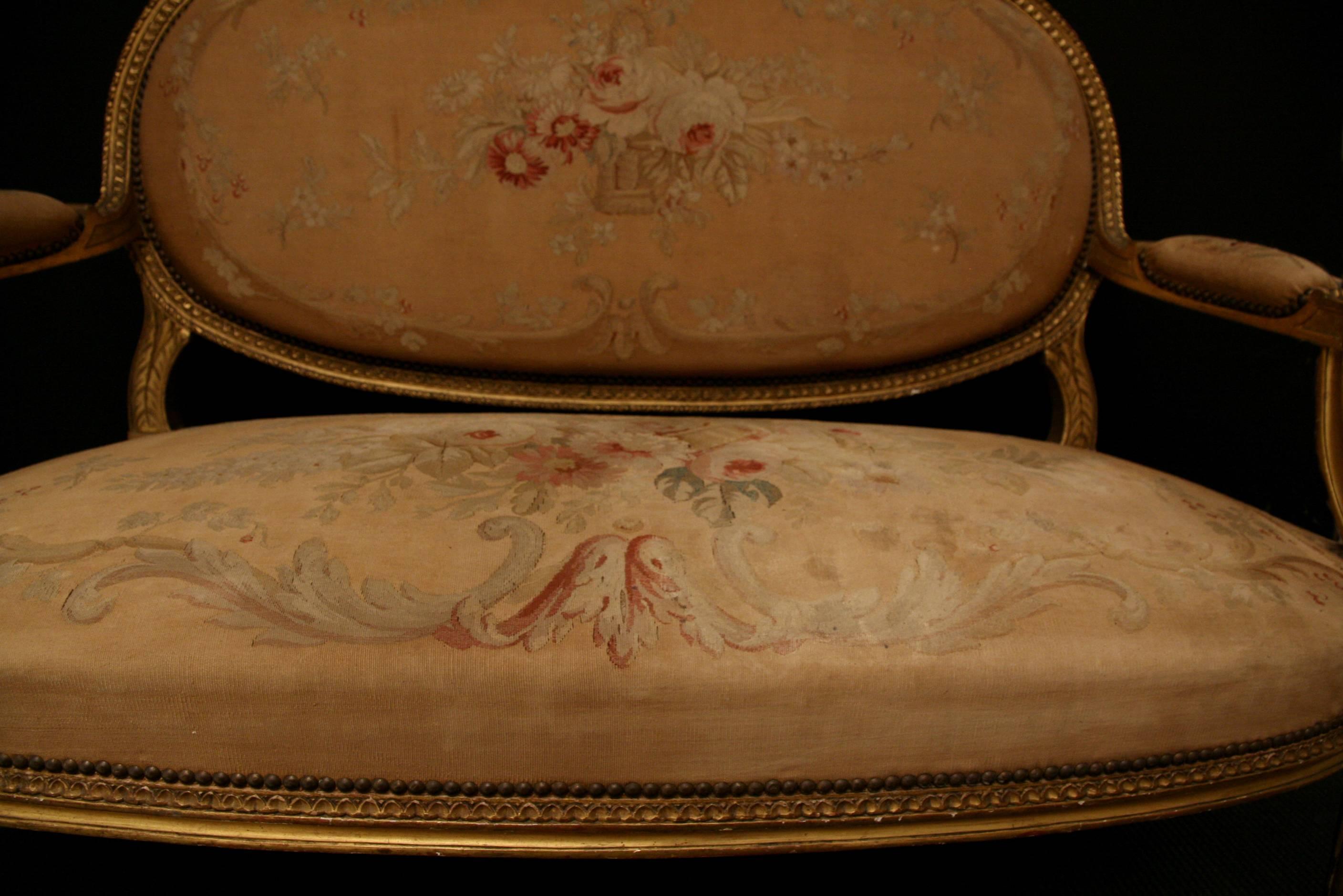 A carved giltwood Louis XVI style settee. The delicately carved floral frames with a ribbon bow crest, Stand on fluted tapering legs, with oval backs and open swept arms, upholstered in Aubusson style tapestry.

Seat height 41cm.