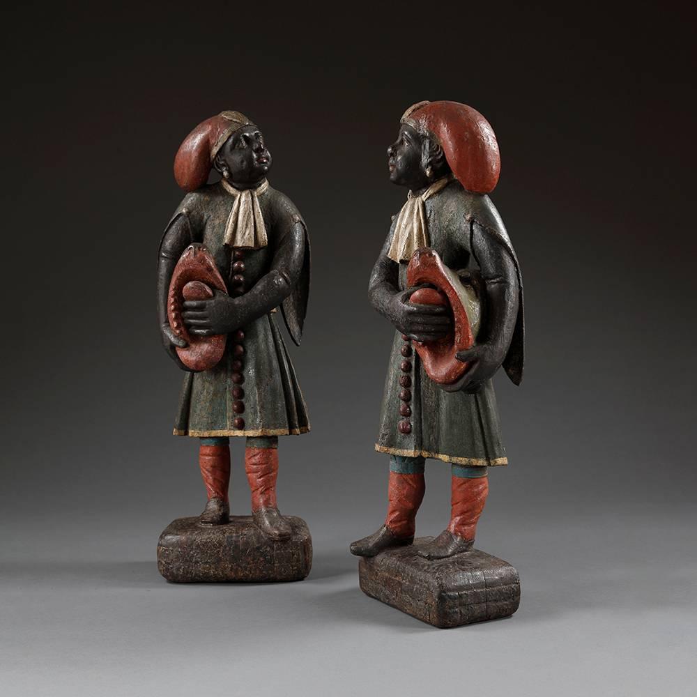 A pair of early 18th century Indian figures delicately carved with red hats, white cravats and holding the tongues of stylised animal with knee length buttoned tunics and red socks standing on black blocked bases. 

These figures would have probably