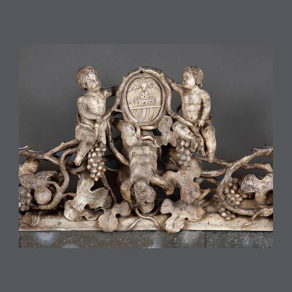 An early 18th century mirror profusely carved with putti and hanging grapes surmounted by a framed crest enclosing the arms of the de Capitani d’Arzago family from Lombardy the whole enclosing an early 19th century plate.