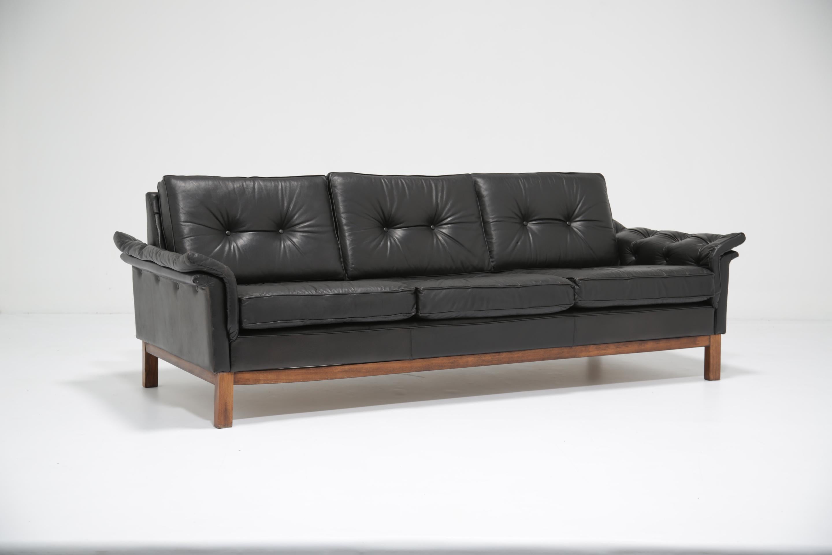 Ce sculptural canapé trois places des années 1960 capture le confort raffiné du design scandinave du milieu du siècle. Recouvert de cuir noir, il présente de profonds coussins de dossier boutonnés et de larges accoudoirs doucement paddés qui