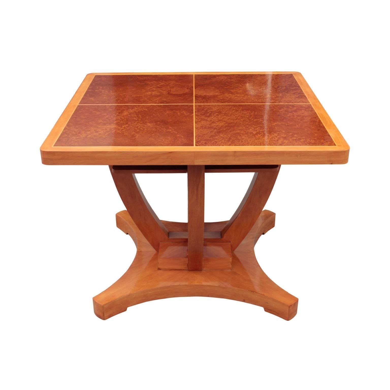Fine table in maple featuring square top with rounded corners and four panels in bird’s-eye maple bordered with inlay banding in satinwood; raised on four elegant curved supports and a cruciform and four blocks plinth; resting on quadripartite base