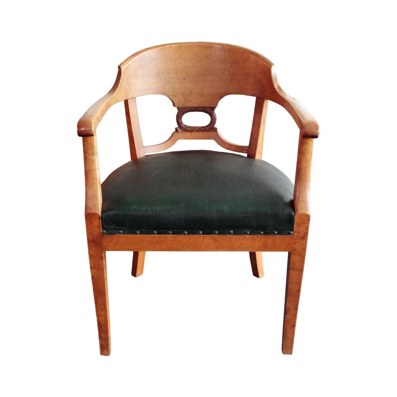 Elegant desk armchair featuring fan shaped barrel backrest decorated with laurel leaf carving in beech; arm rests ending in subtle scrolls; saber legs. Seat with preserved dark green leather with nailheads. In Karelian birch, flame birch and beech.