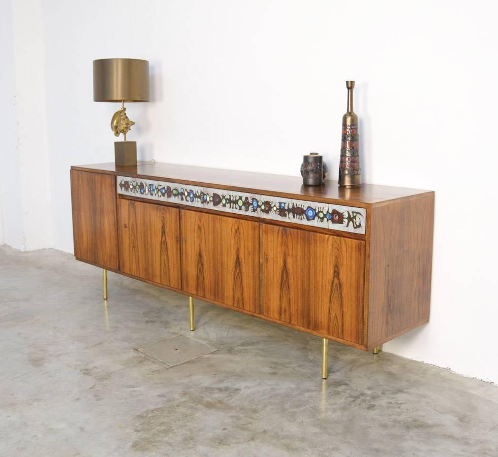 This dark rosewood sideboard was designed by Alfred Hendrickx for Belform in the 1950s.
The famous ceramist Achiel M. Pauwels created the tiles which cover the 3 drawers. This unique sideboard has a bar with light on the left side.
The feet are