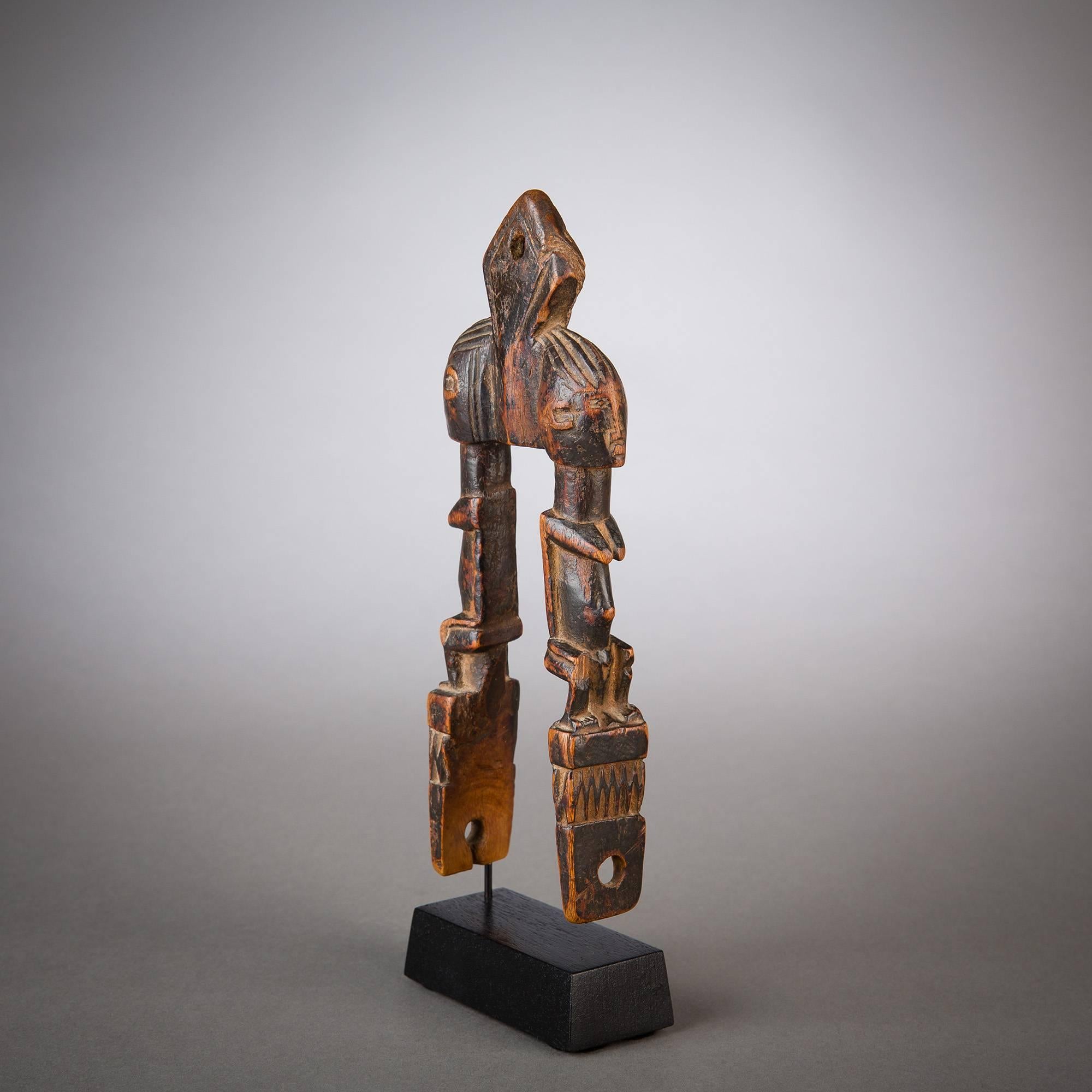 Delicate symmetry defines this old, rare heddle pulley, which features twin figures posed back to back across an open space. The slim, caliper like silhouette of the pulley has been shaped with delicacy, and the dark patina shows lovely reddish