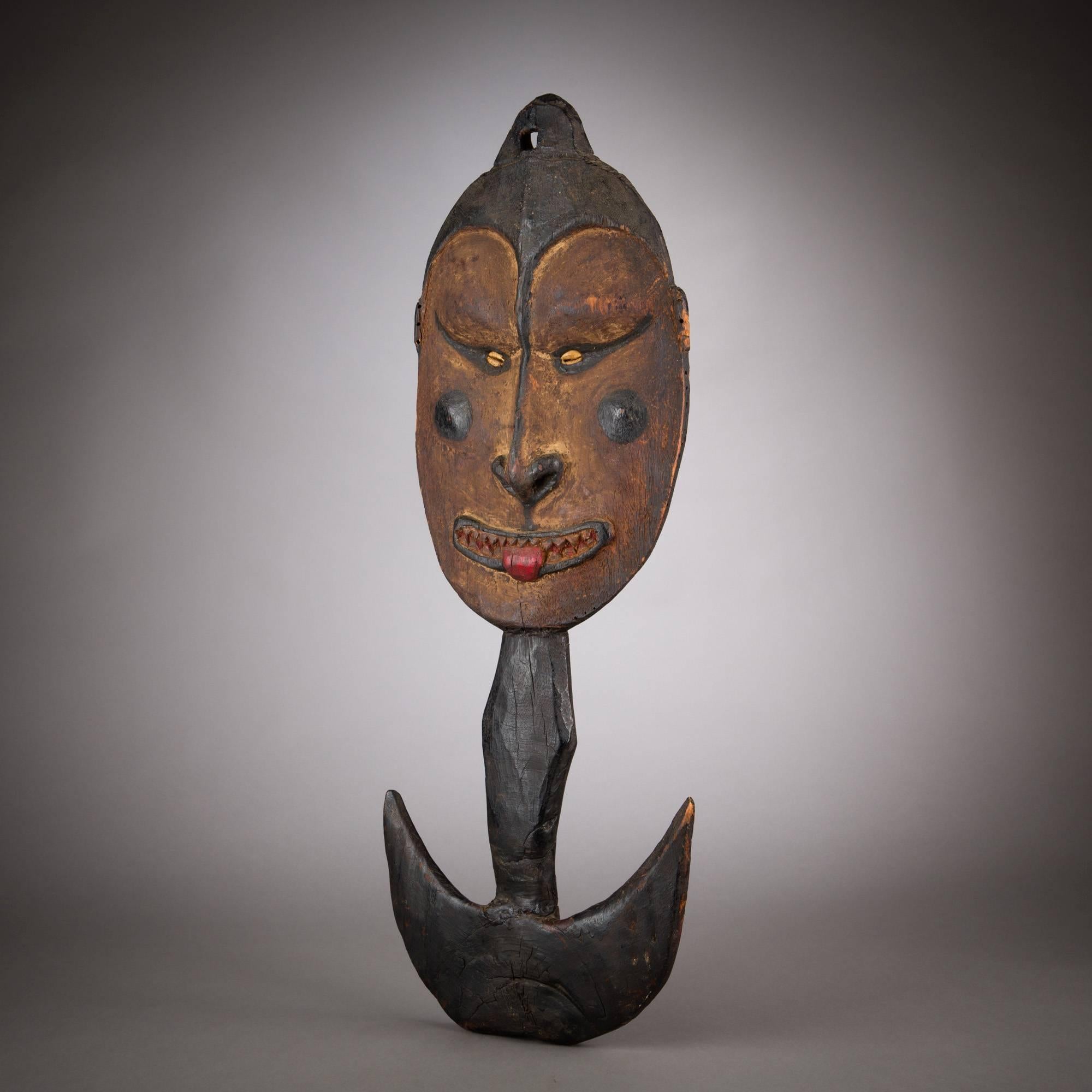 A compelling suspension hook with large, defined face. Into features of textured umber are set cowry shell eyes, a mouth with jagged teeth, projecting tongue and an arrow-shaped nose that plunges from a dark brow. The darkened cheeks cleverly act as