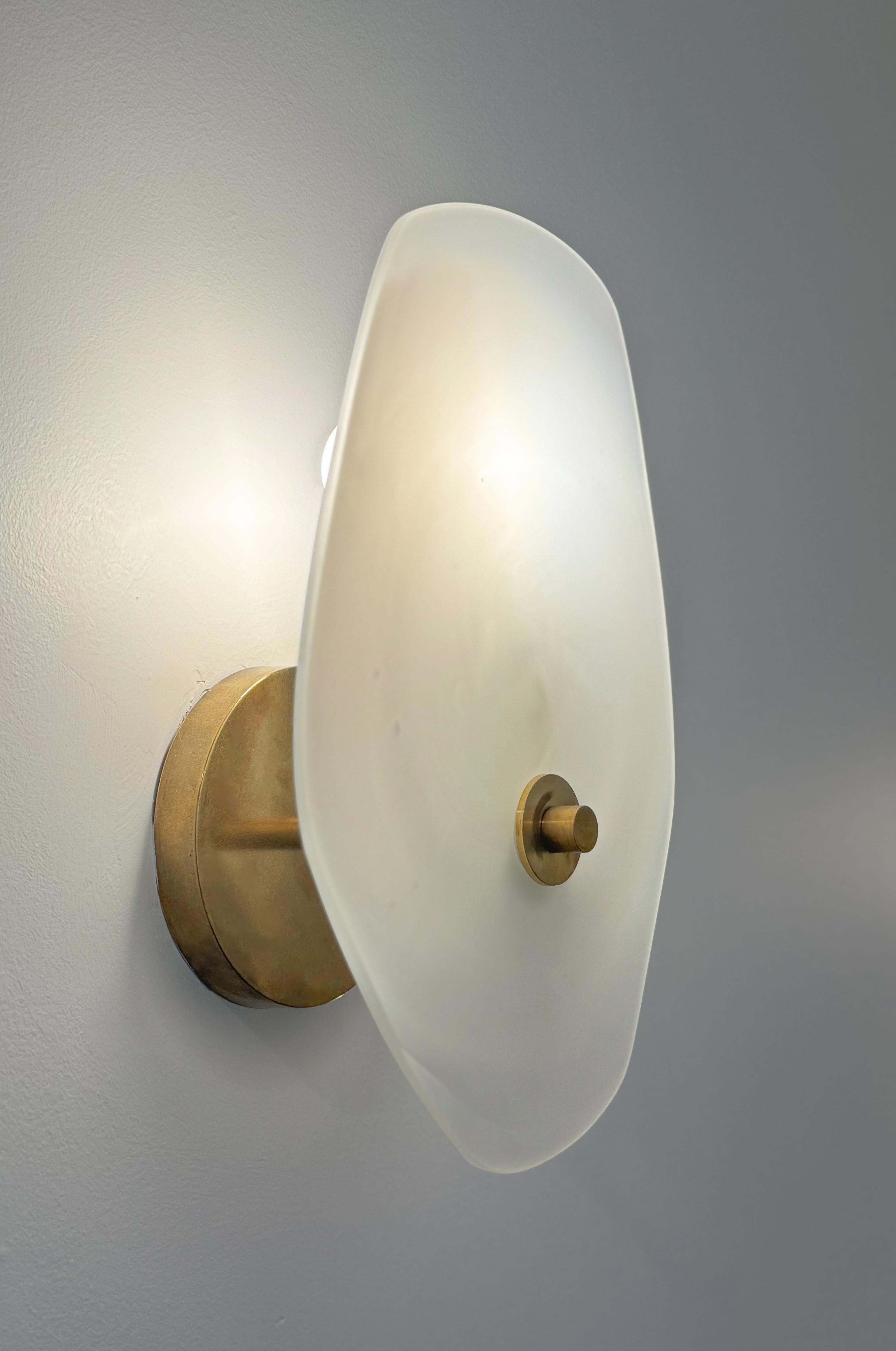 The Clypea Sconce by Brent Warr is a refined study in balance and materiality. Featuring a hand-blown tinted glass disc paired with a warm brass armature, this modern wall sconce offers both sculptural elegance and soft illumination. The minimal