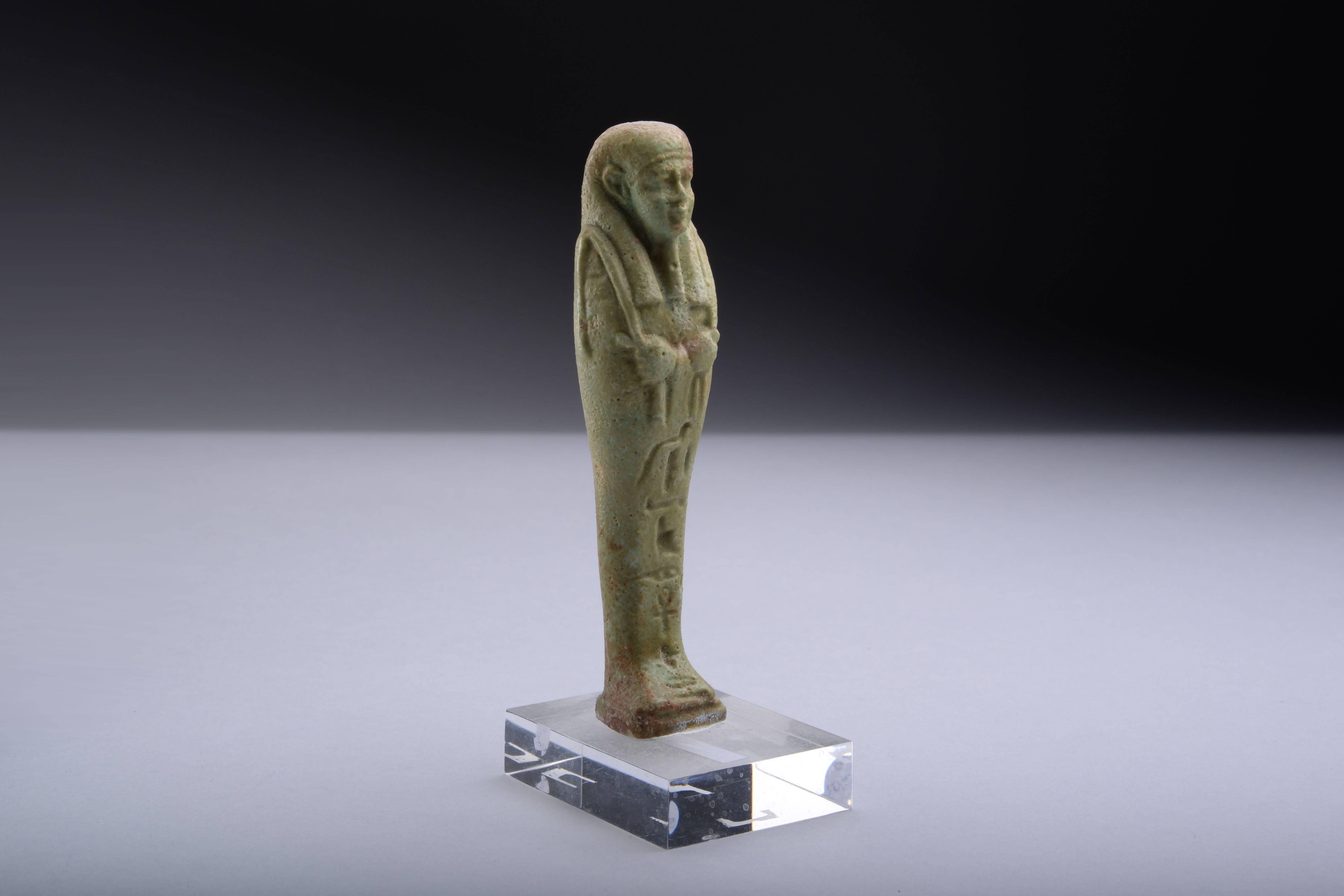 A quality, serene, ancient Egyptian faience shabti for Ankh-Her, dating to the Late Period, circa 664 - 332 BC.
 
The figure is depicted mummiform, with arms crossed holding two hoes, wearing tripartite wig and false beard, a vertical line of