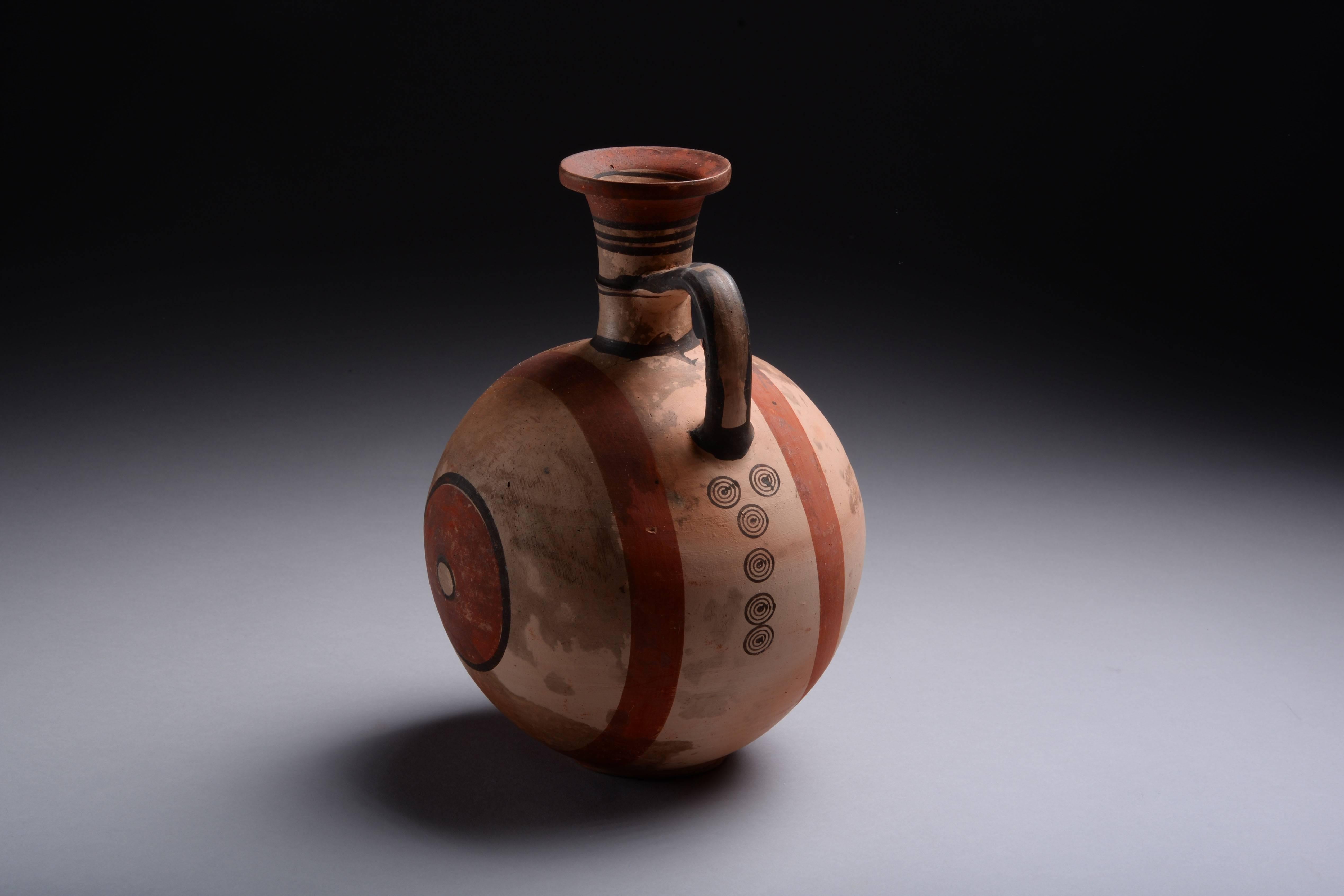 An ancient Cypriot iron age geometric period oinochoe (or wine jug), dating to circa 750-650 BC.

Standing on a ring base with a large, round body, cylindrical flaring neck, splayed mouth and loop handle. Decorated throughout with bands and