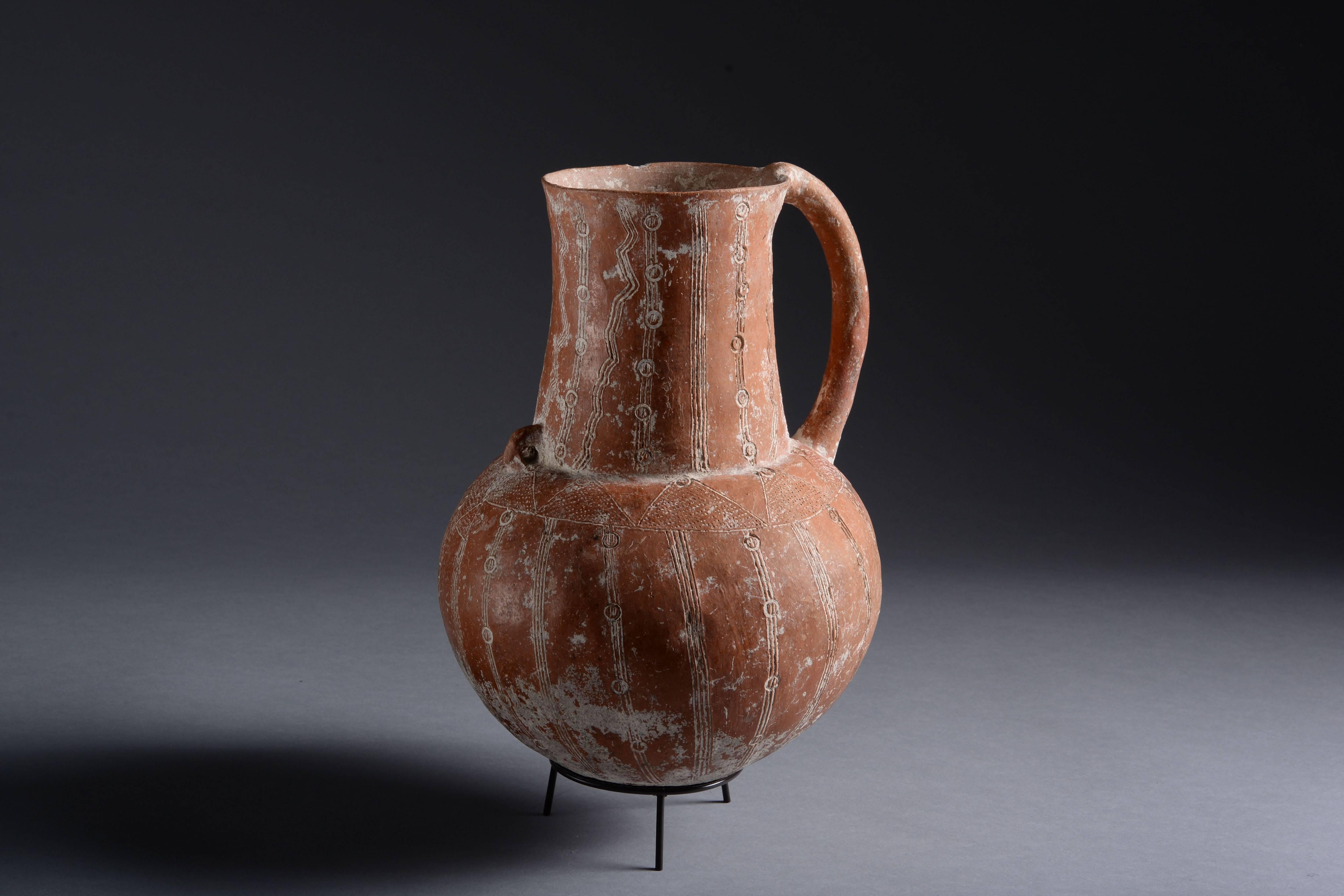 An early bronze age cypriot jug, dating to approximately 2400-1900 BC. Of typical red burnished pottery.

The globular body with wide cylindrical neck. Beautiful incised decorations throughout, lime filled and displaying a combination of zig-zags,