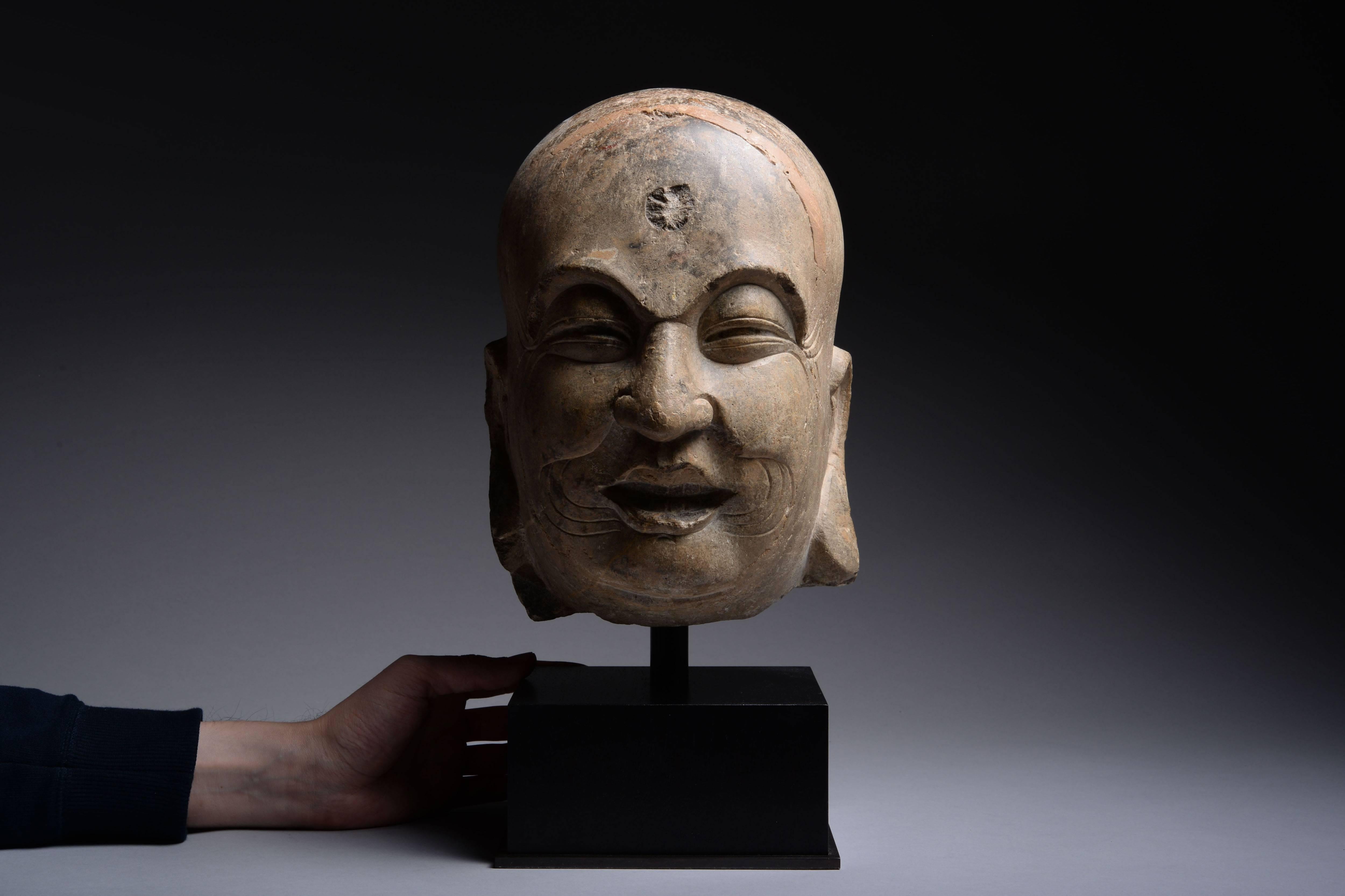 An ancient Chinese limestone sculpture depicting the head of a laughing lohan. Dating to China's Song Dynasty, circa 960 AD.

The lohan is shown with typical chubby face and bald head, his smiling mouth with teeth showing, the prominent creases in