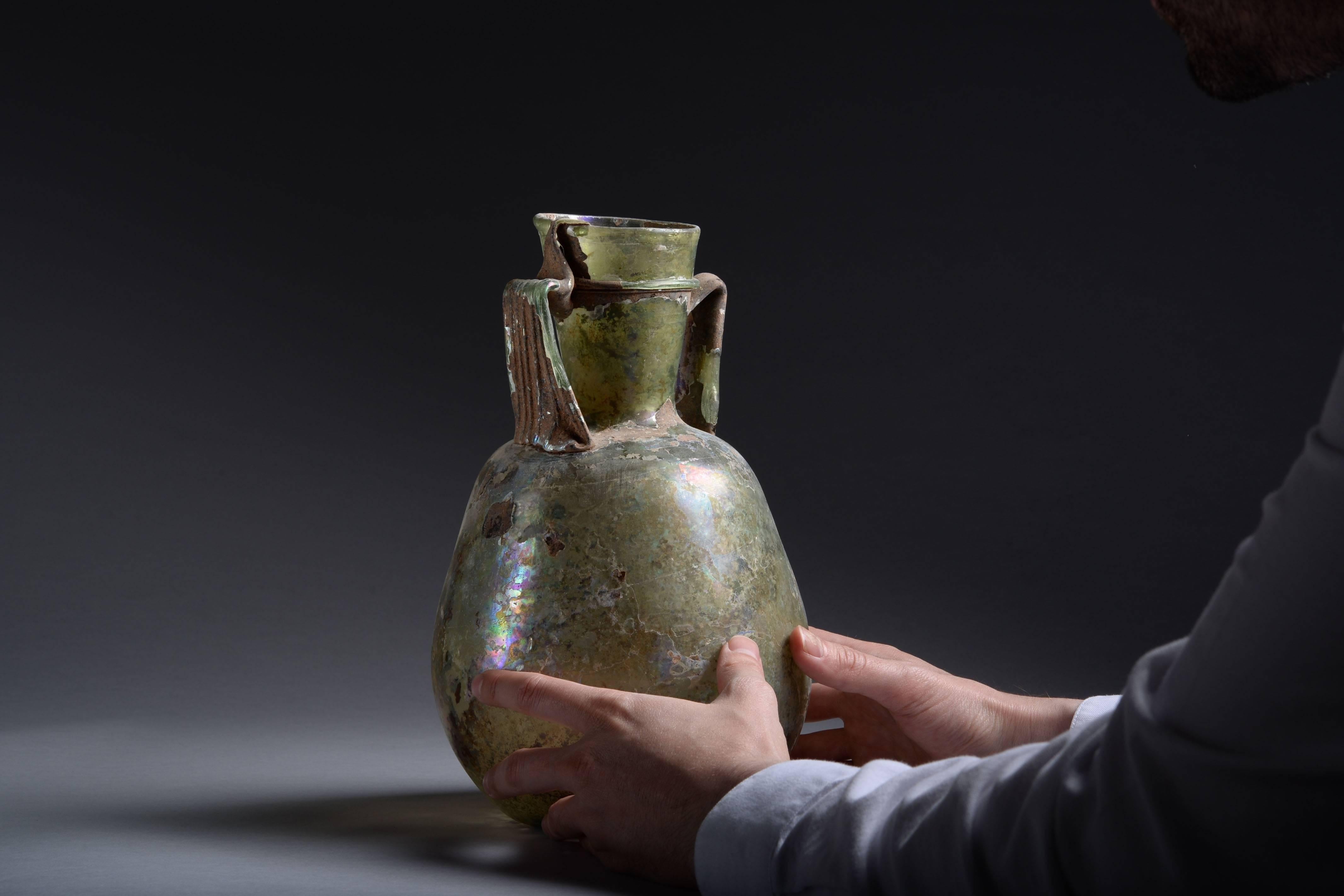 A large ancient Roman green glass Amphora from the Near East, dating to the 3rd century AD.

Free blown and sitting on a slightly indented base, with elongated body, rounded shoulders and flaring cylindrical mouth. The vessel is adorned with two