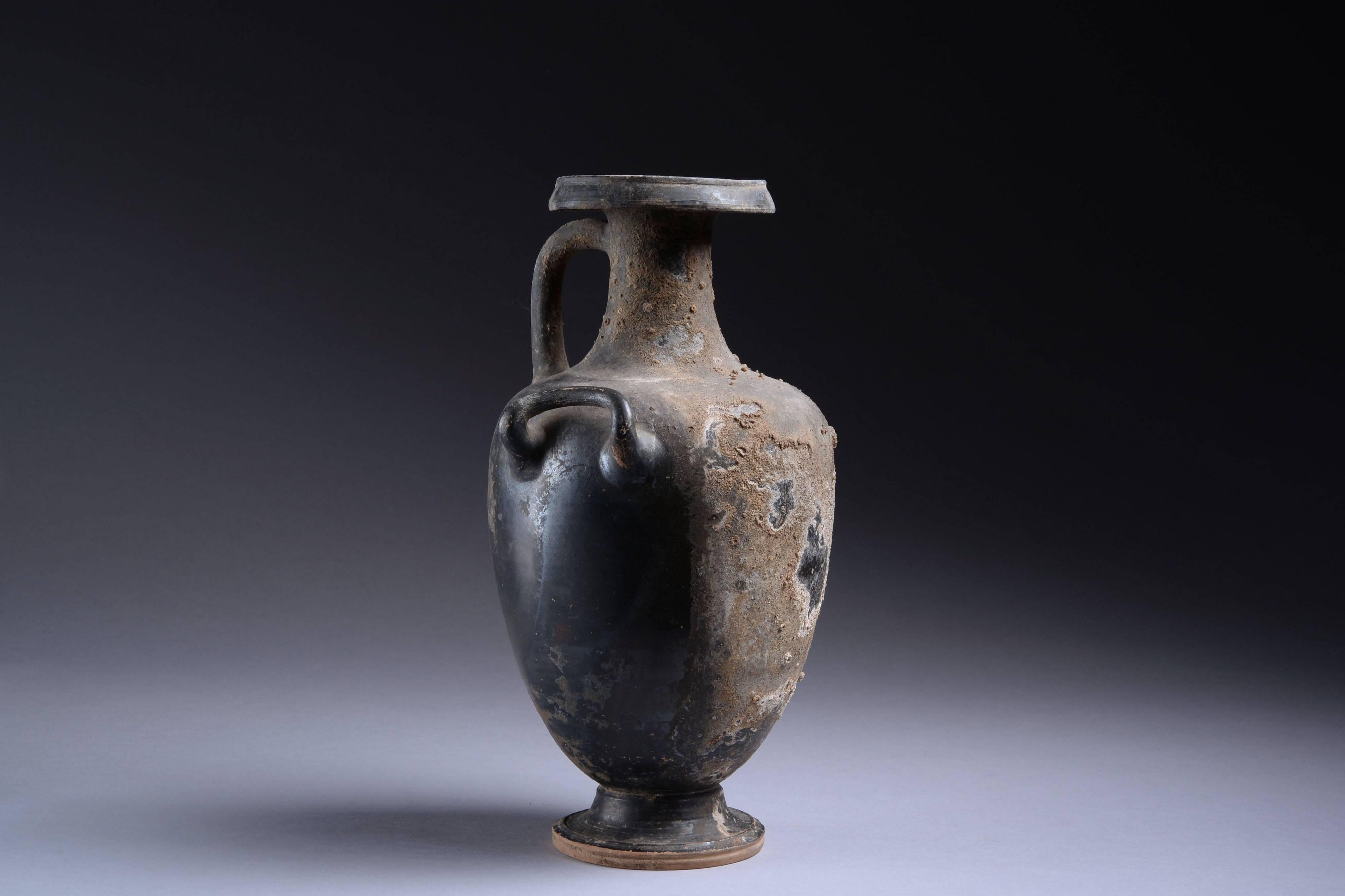A large, elegant ancient Greek blackware hydria, in exceptional condition, dating to the 4th century BC.

The piece stands on a circular base, with acorn shaped body, cylindrical neck and splayed mouth. A large loop handle arches from the neck to