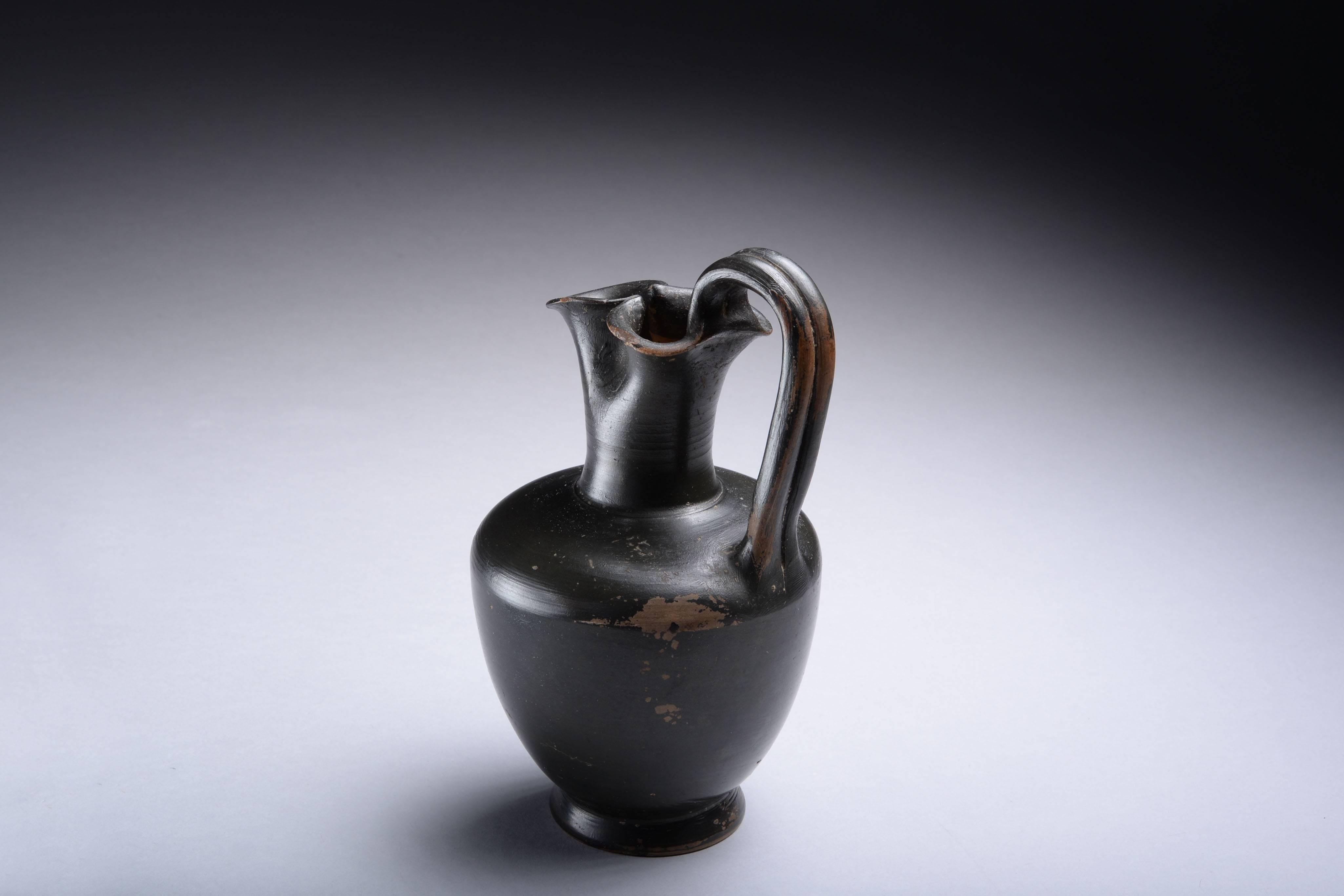 A well provenanced ancient Greek blackware oinochoe jug, dating to the 4th century BC.

The piece stands on a circular base, with acorn shaped body, cylindrical neck with trefoil mouth, and elegant, double banded loop handle. Covered with a