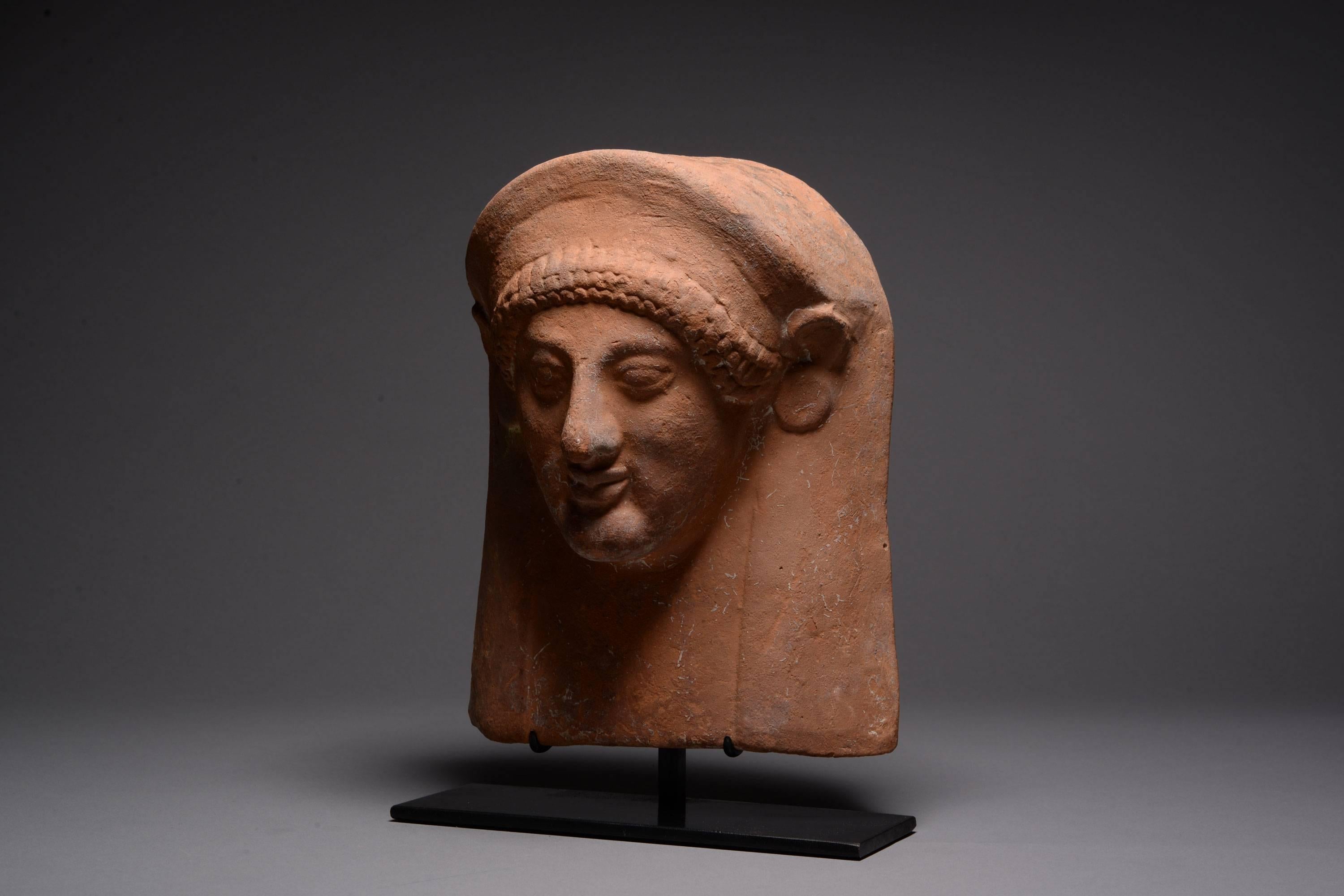 An ancient Greek terracotta female votive mask, or protome, dating circa 550 BC.

Of striking Greek archaic style, showing a female figure wearing disk earrings, a stephane and a himation pulled up over the top of her head. The hair emerging from