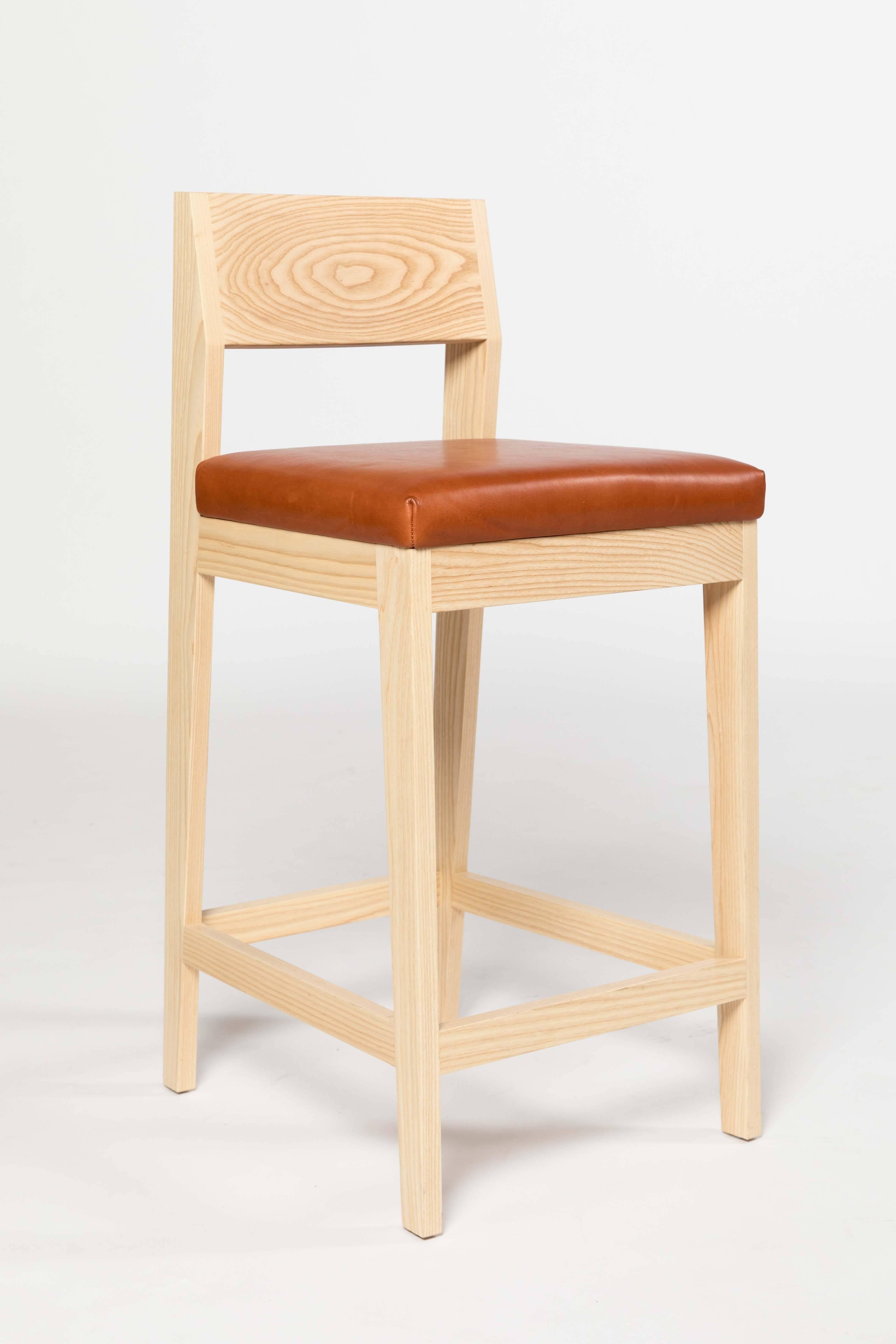 The Shelley stool by Kate Duncan is handcrafted from solid wood. It incorporates clean lines and intersecting angles to maximize comfort. This high stool features a sloped curved back that cradles resting shoulders and a leather seat with integrated