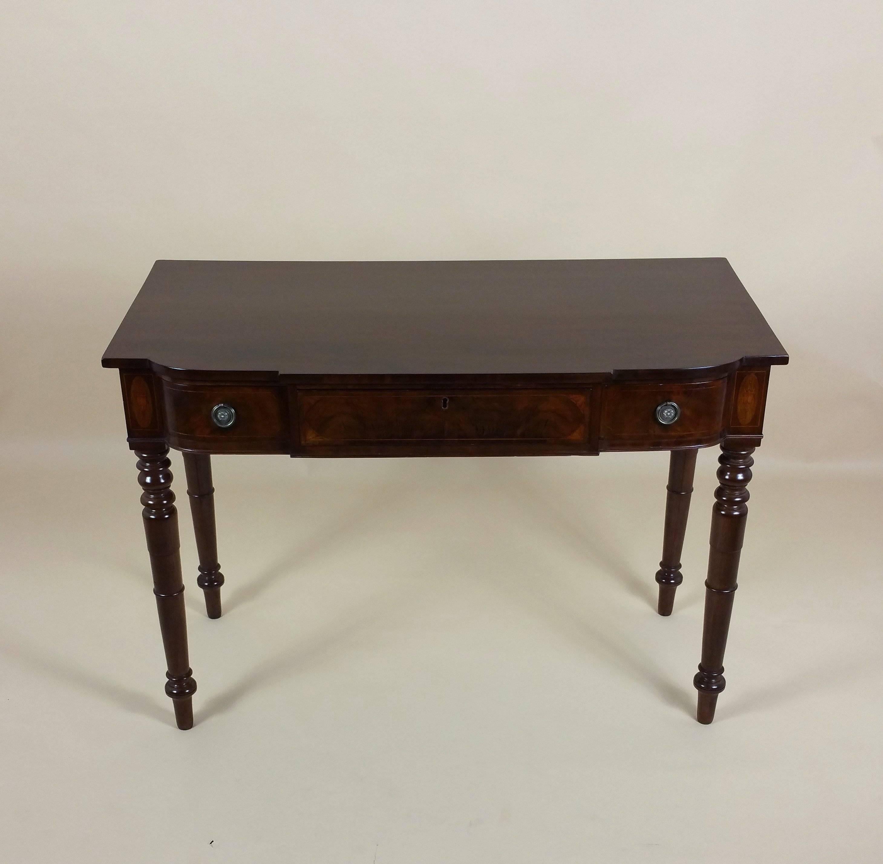 This handsome and well-proportioned George III inlaid mahogany breakfront side table features a large central drawer with a smaller curved drawer on either side, finished with brass pull rings. The side table is decorated with an inlaid design of