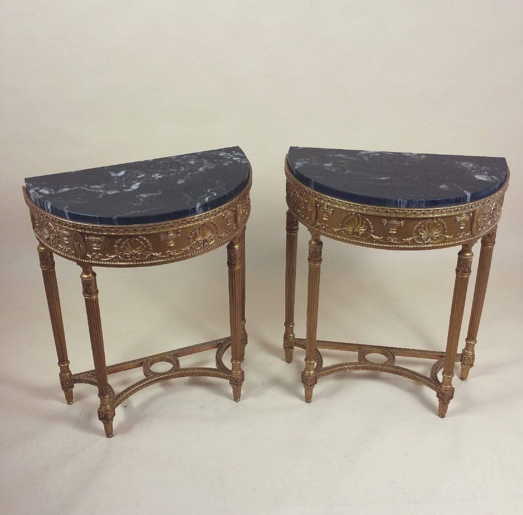 This lovely pair of demilune gilt console tables with marble tops depicts urns and anthemions in the style of Robert Adam, circa 1900. The tables stand on delicately tapered reeded legs with a beveled edge around both marble tops. The shaped