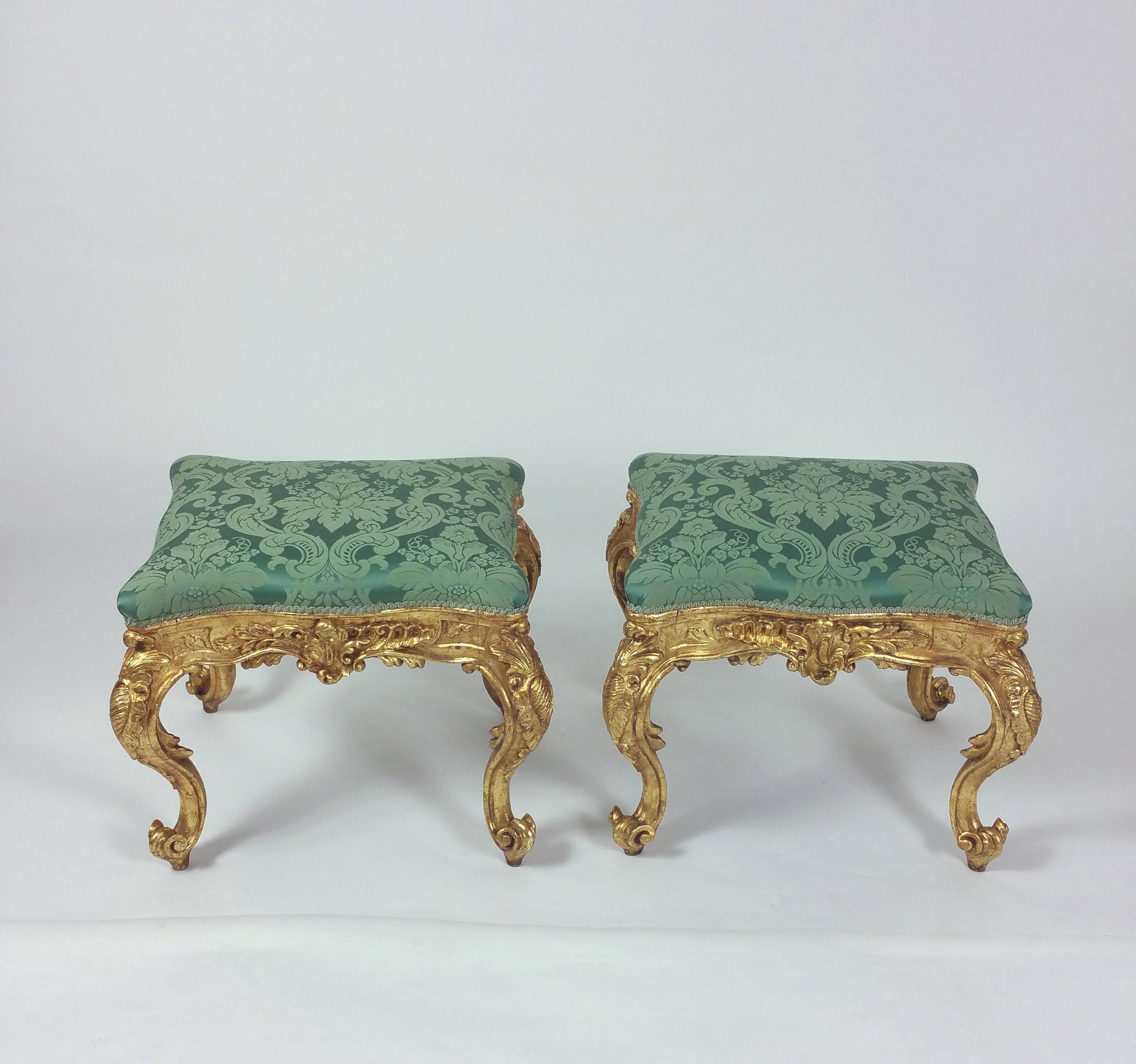 These elegant and fine French ornately carved giltwood stools are serpentine in form. They feature cabriole legs on scroll feet decorated with scrolls and acanthus leaves, and are upholstered in a green damask fabric. Each stool measures 24 in – 61