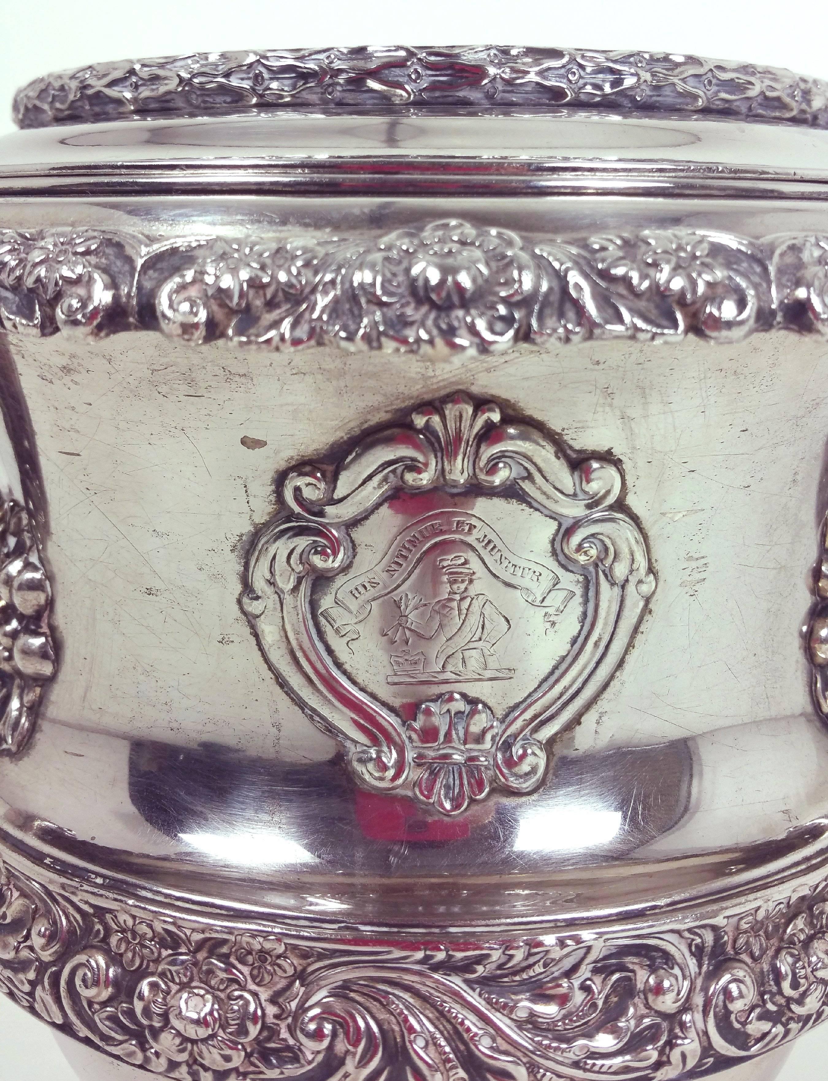 This handsome and ornately detailed 19th century silver plated champagne cooler features a twin handle design with a heraldic cartouche. The cooler has several bordering trims of acanthus leaves, flowers and fruit, as well as a top rim which is