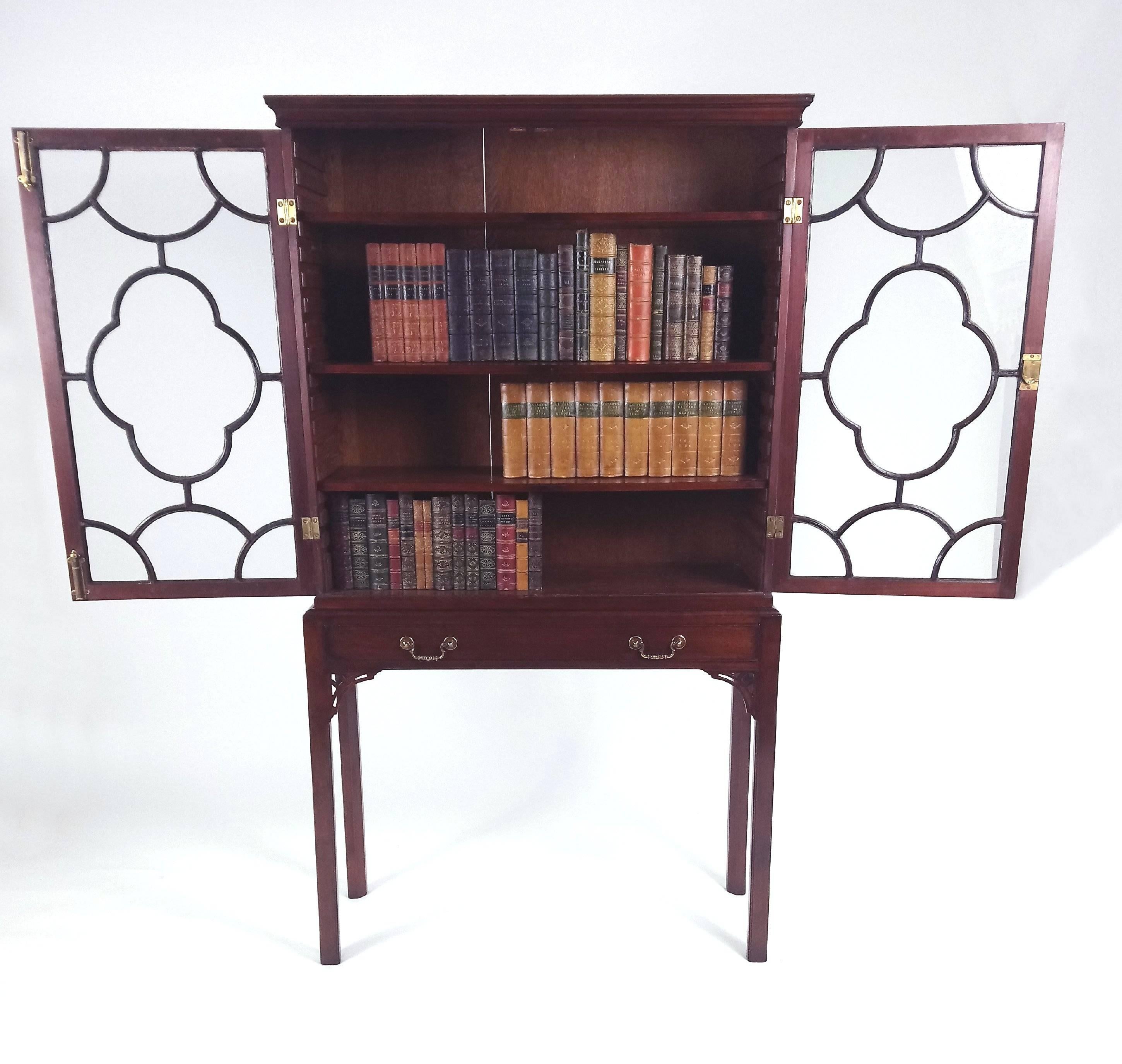 This charming and highly desired size George III mahogany two astragal glazed door bookcase features adjustable shelves and a single central drawer on stand with the original brass handles and pierced winged brackets on square chamfered supports.