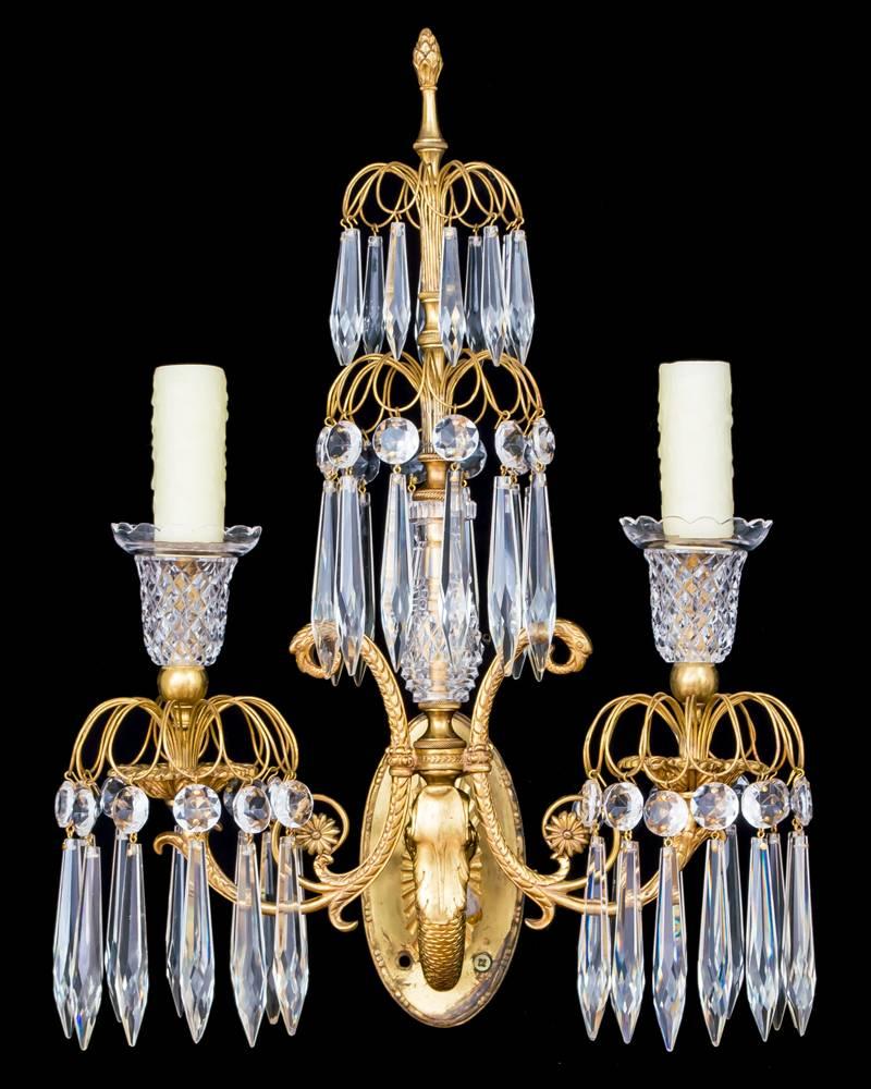 A pair of gilt dolphin wall lights supporting two serpentine j arms issuing diamond cut candle nozzles and drop hung sprays the central cut glass columns also with drop hung sprays.