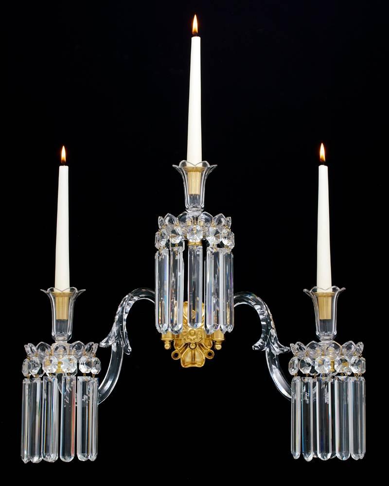 A pair of English William IV three-light ormolu-mounted cut-glass wall lights the ormolu back plate surmounted by three-leaf cut candle arms with flute cut nozzles and drip pans hung with rule drops.