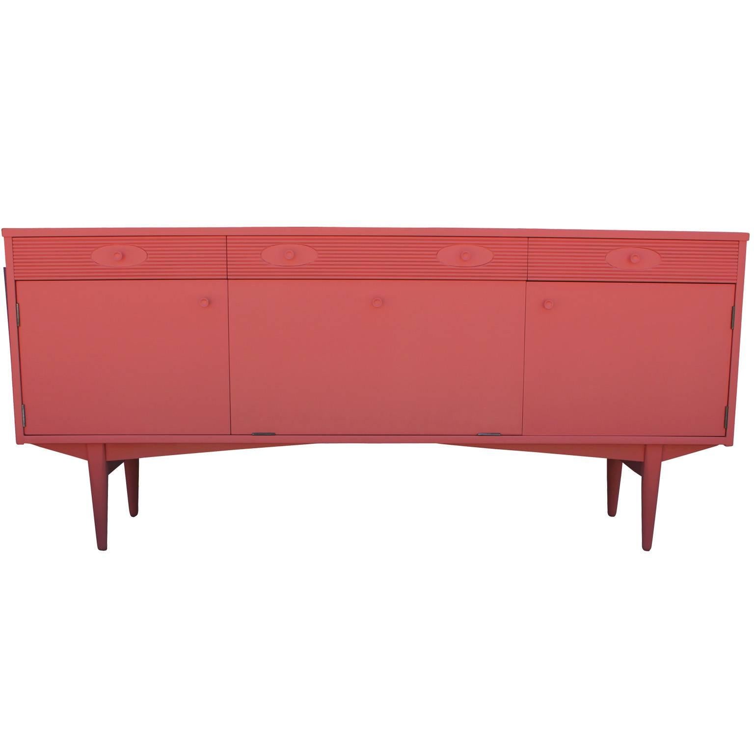 Charming sideboard finished in an ultra-smooth and glossy coral pink lacquer. Cabinet doors in the left and right and a center drop down door all open to reveal a single shelf. Three pull out drawers provide additional storage.