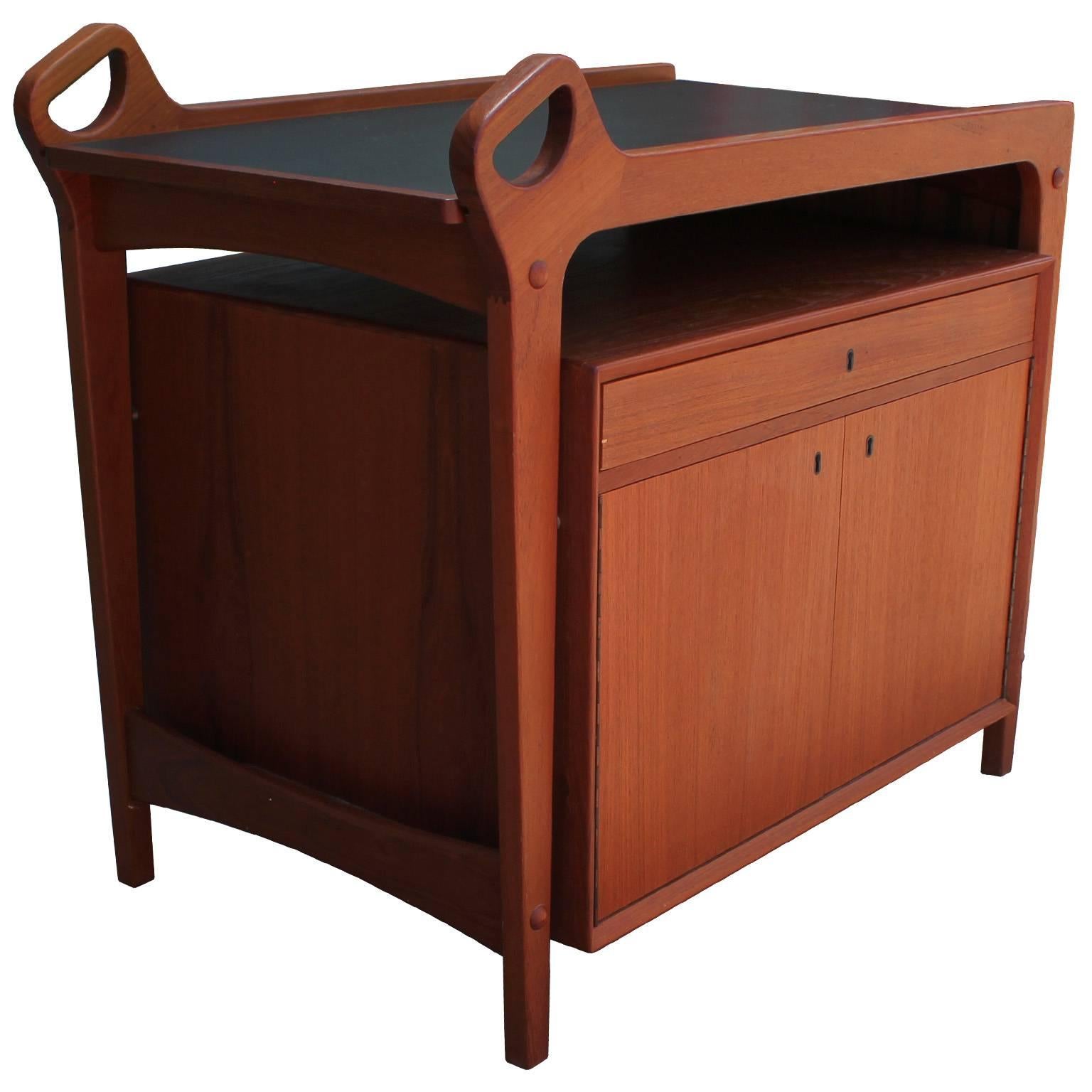 Sculptural Danish bar cart in teak with a black laminate surface. Cart has excellent lines with a wonderful use of negative space. Serving table top slides into place an extends. A single drawer is slotted and lined in green felt. Two cabinet doors
