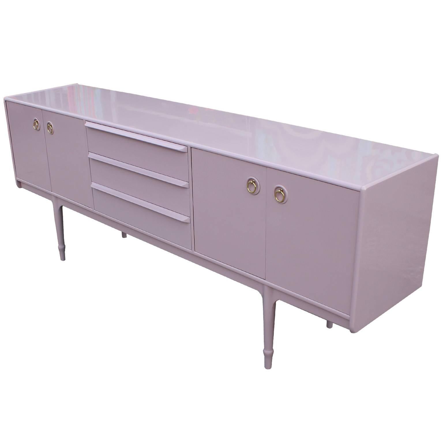 Fabulous sideboard or credenza by McIntosh. Sideboard is freshly finished in an ultra smooth pale pink or lavender lacquer. Brass ring pulls complete the piece. Cabinet doors on either side open to a single shelf. Three drawers provide additional