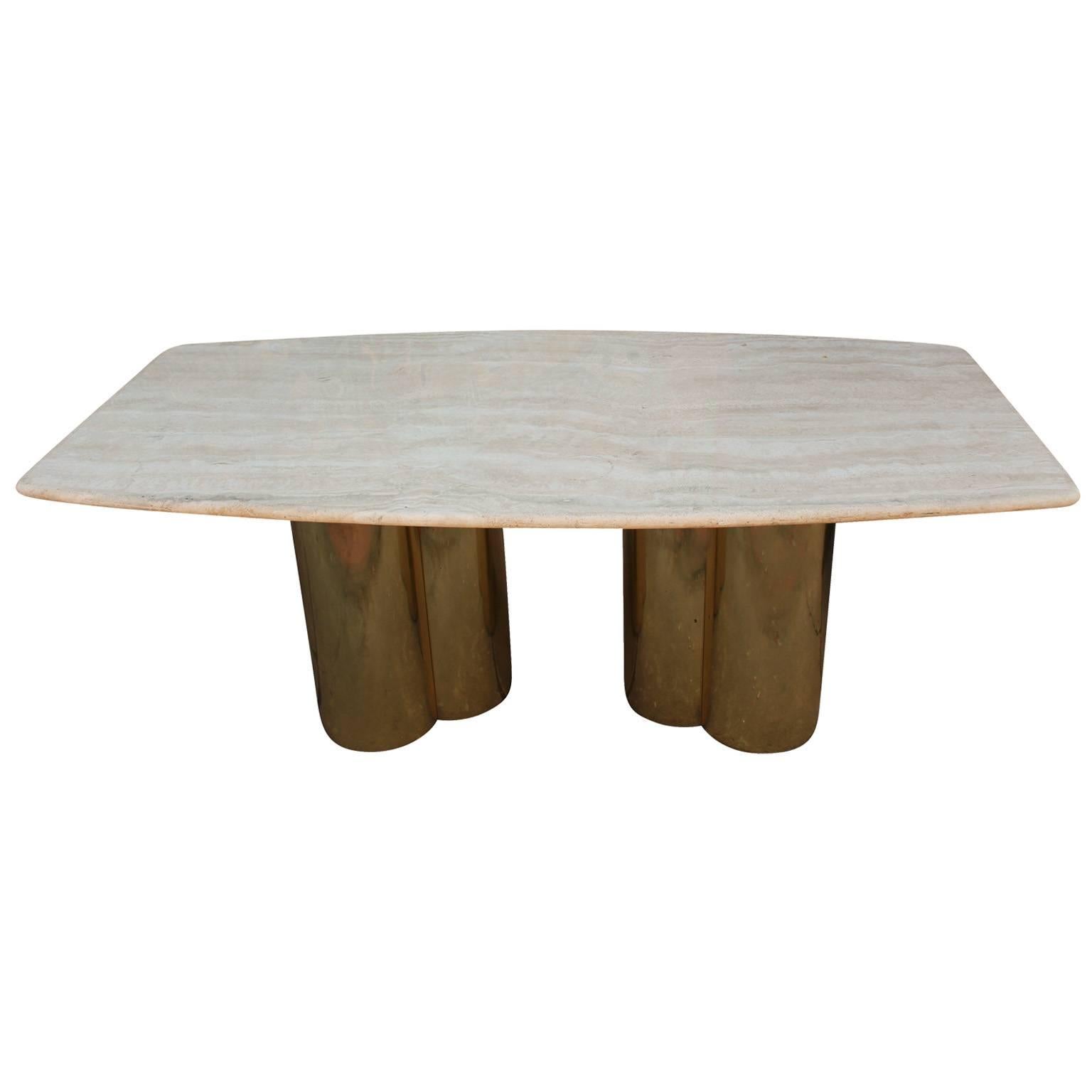 Luxe travertine topped dining table. Boat shaped travertine top has a lovely grain. Top rests upon two shiny brass, trefoil shaped bases. The perfect hollywood regency style dining table. The brass pedestals are believe to be made by Curtis Jere.