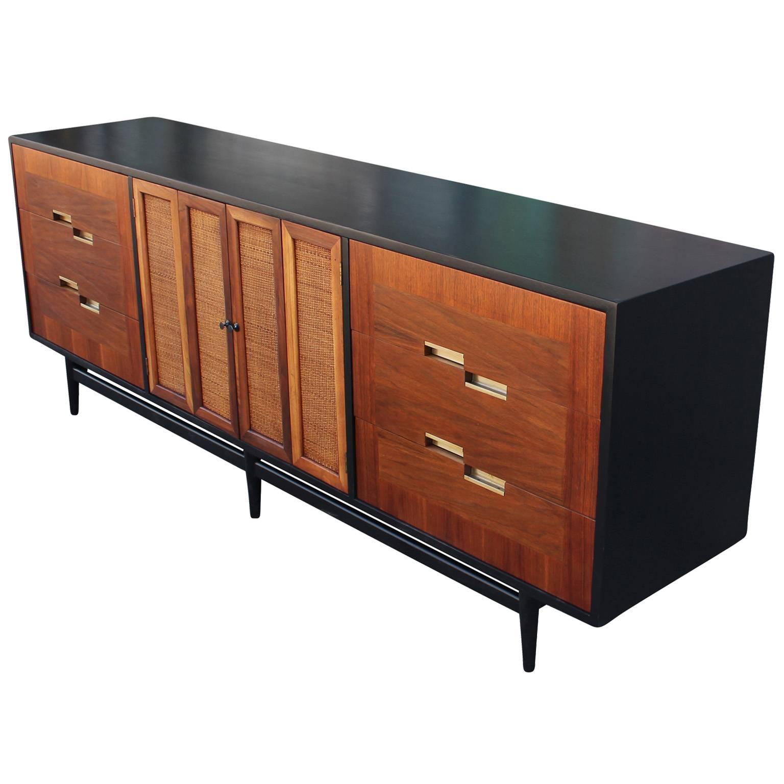 Excellent long and low dresser by American of Martinsville. Case is finished in ebony and drawer fronts in a medium walnut. Three drawers with alternating brass hardware on either side. Raffia front bi-fold doors open to three drawers. An airy base