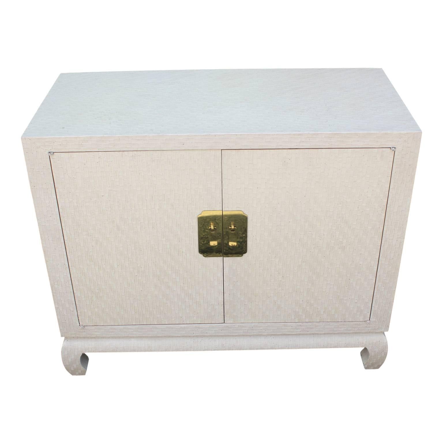 Cream colored lacquered grass cloth cabinet from Baker's Far East collection. Ming style turned legs, and large brass escutcheons backing solid brass drop pulls. The two doors open to reveal a single shelf.