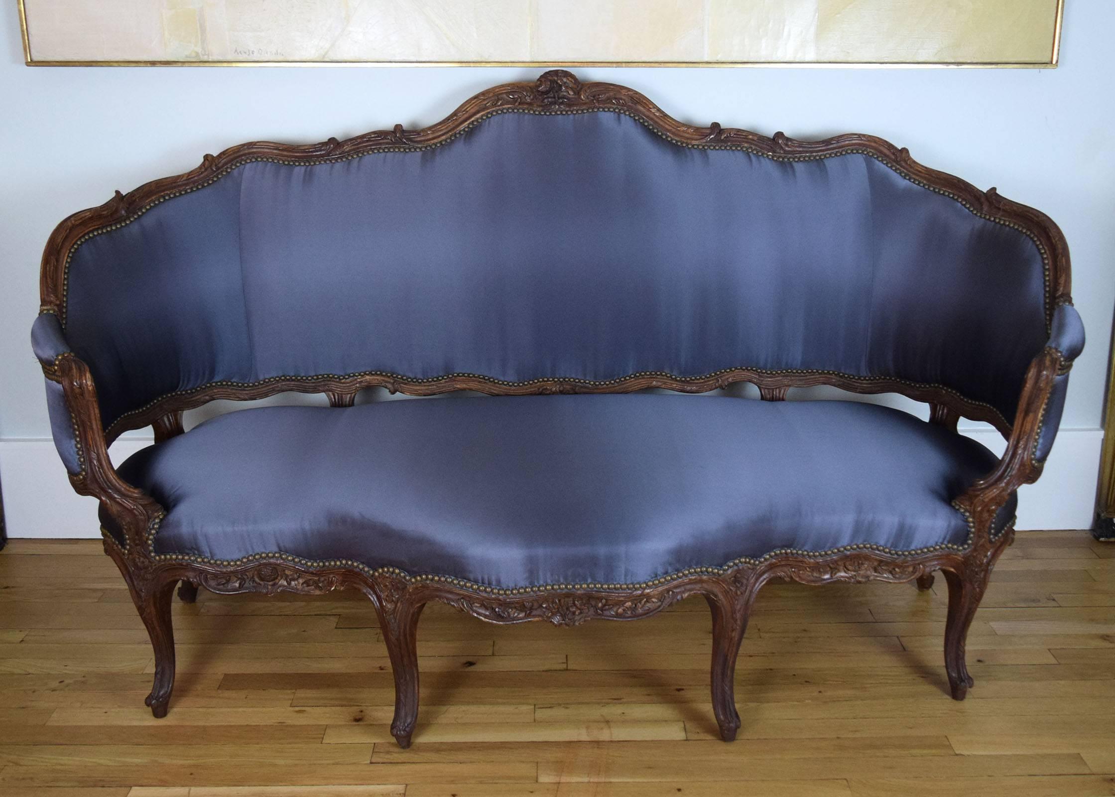 The walnut frame of this large, Louis XV-period sofa is unusual for its highly animated form. It is finely carved with flowers, leaves, bound reeds and scrolling vegetal forms. We have had it reupholstered in slate-blue silk satin.