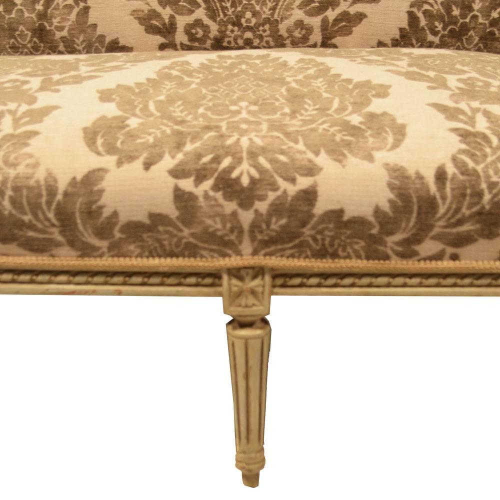 Louis XVI Winged Loveseat featuring an antiqued painted finish in cream with a taupe rub. The loveseat has been newly upholstered in a Schumacher burned velvet fabric featuring an Ikat pattern in cream and beige. Legs are very sturdy, seat depth is