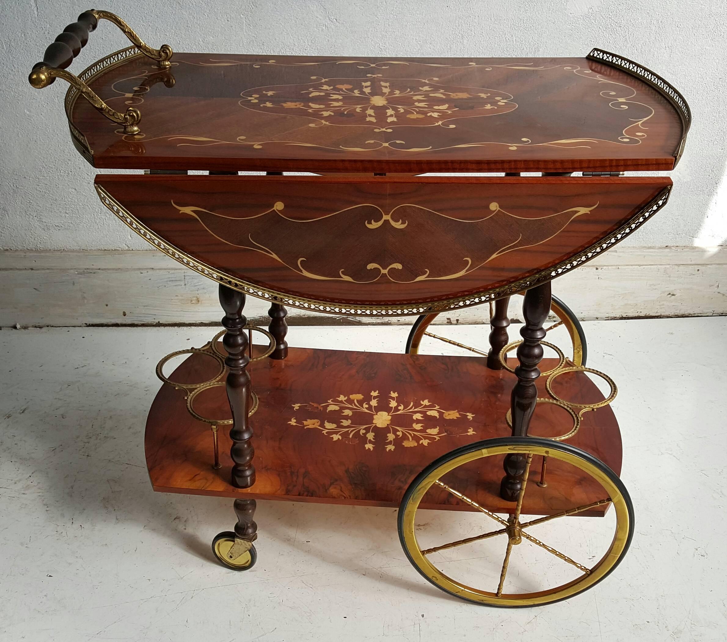 This beautiful vintage Italian drop-leaf bar cart, produced, circa 1950s, includes two tiers of lacquered burl, each inlaid with central floral marquetry, with brass trim to the top gallery and wood handle, the six bottle holders below, as well as