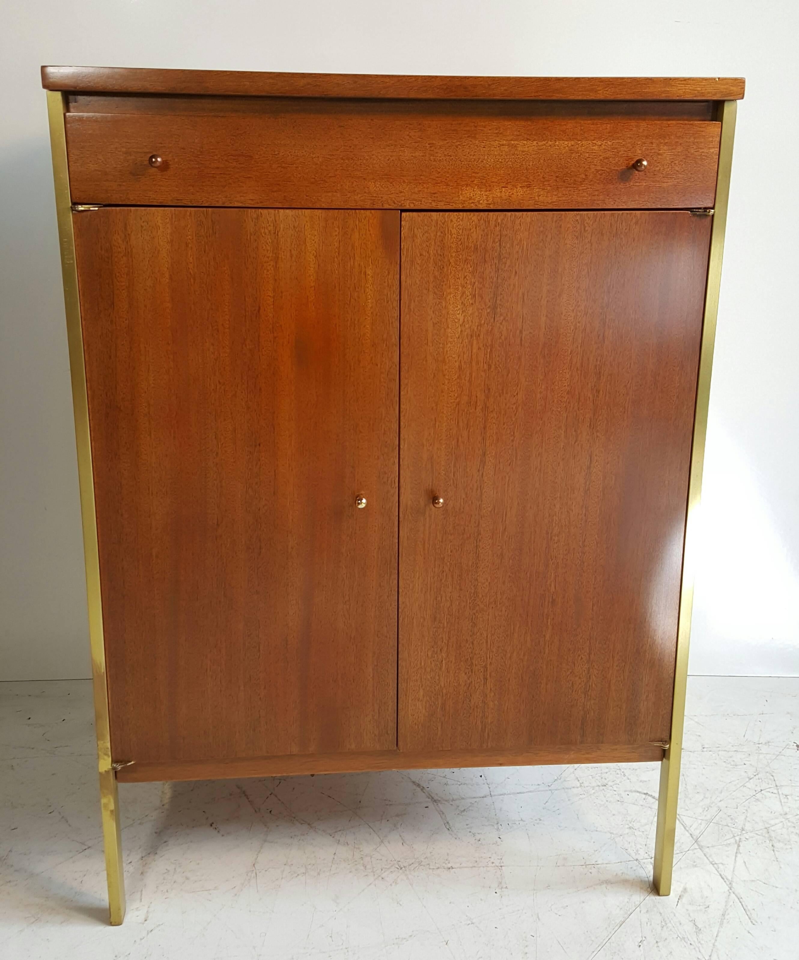 This small server or bar cabinet by Paul McCobb is walnut-stained mahogany with brass frame and pulls. One-drawer, two-doors inside, an adjustable shelf. Great scale and proportion. Retains original Paul McCobb connoisseur label.