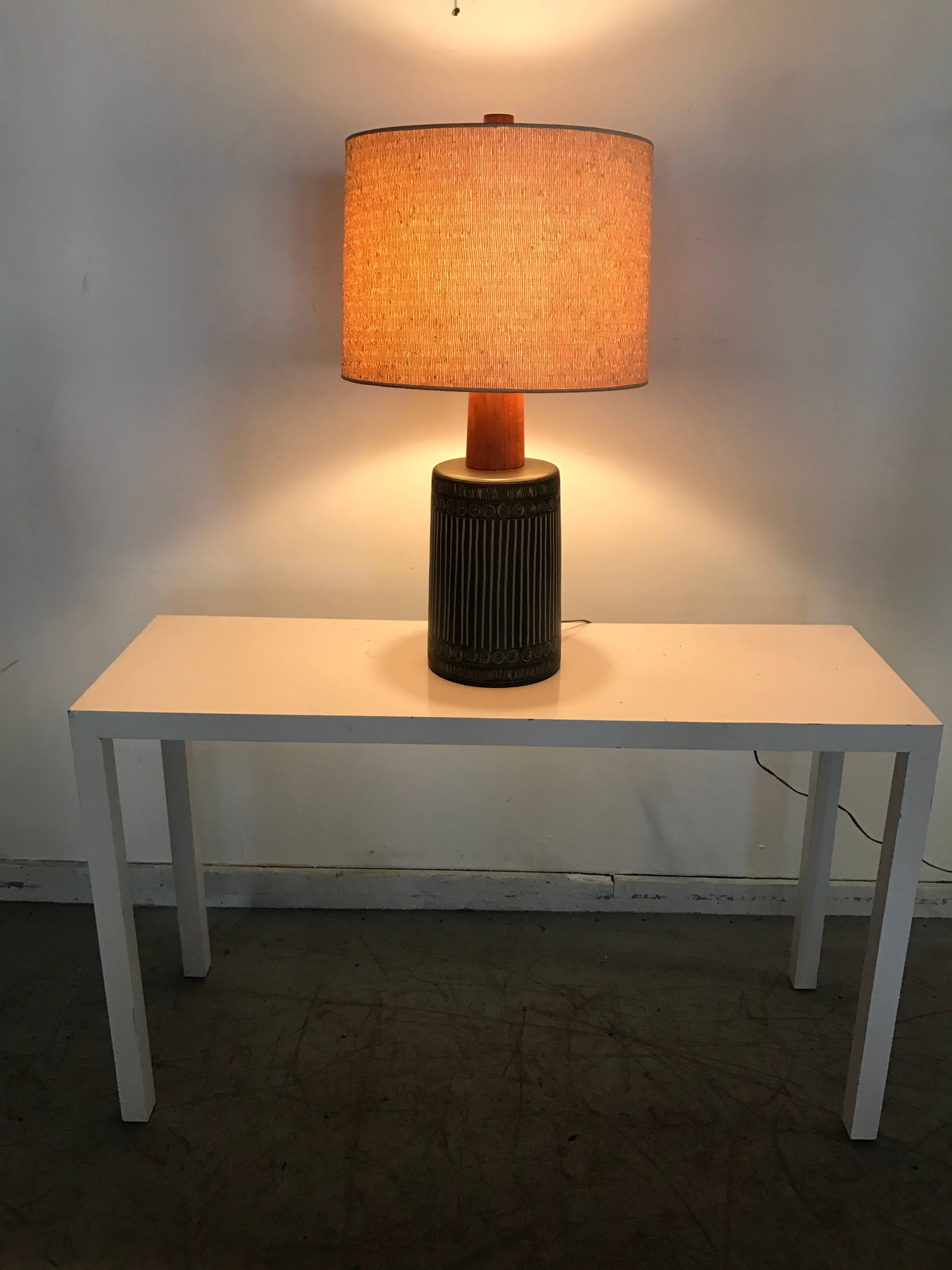 A circa 1960s table lamp, produced for Marshall Studios and signed Martz, retains original teak finial and stem, mounted on glazed grey/black ceramic body, of cylinder form with geometric incising ,retains original lamp shade, Markings include