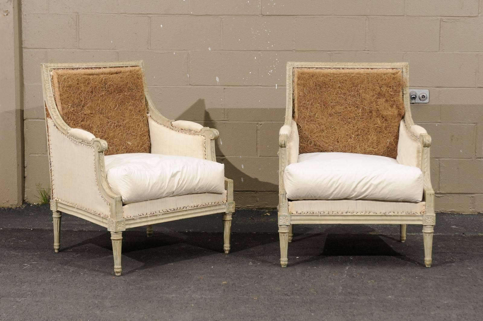 A pair of French early 20th century Louis XVI style painted bergère chairs from Southern France. These French chairs feature a linear back over partially upholstered and scrolled arms. Four carved rosettes adorn the knees of these French chairs,