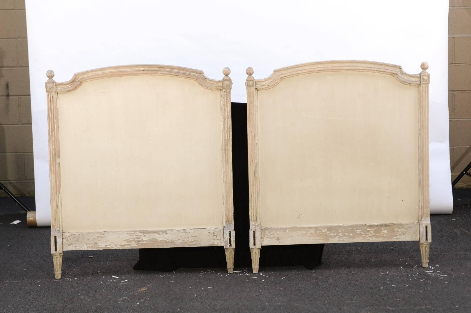 This pair of French 19th century twin-sized bed headboards features elegant Louis XVI style frames with round finials, carved rosettes over nicely fluted side posts. The central wooden panel is painted in a off-white / light cream tone. The ensemble