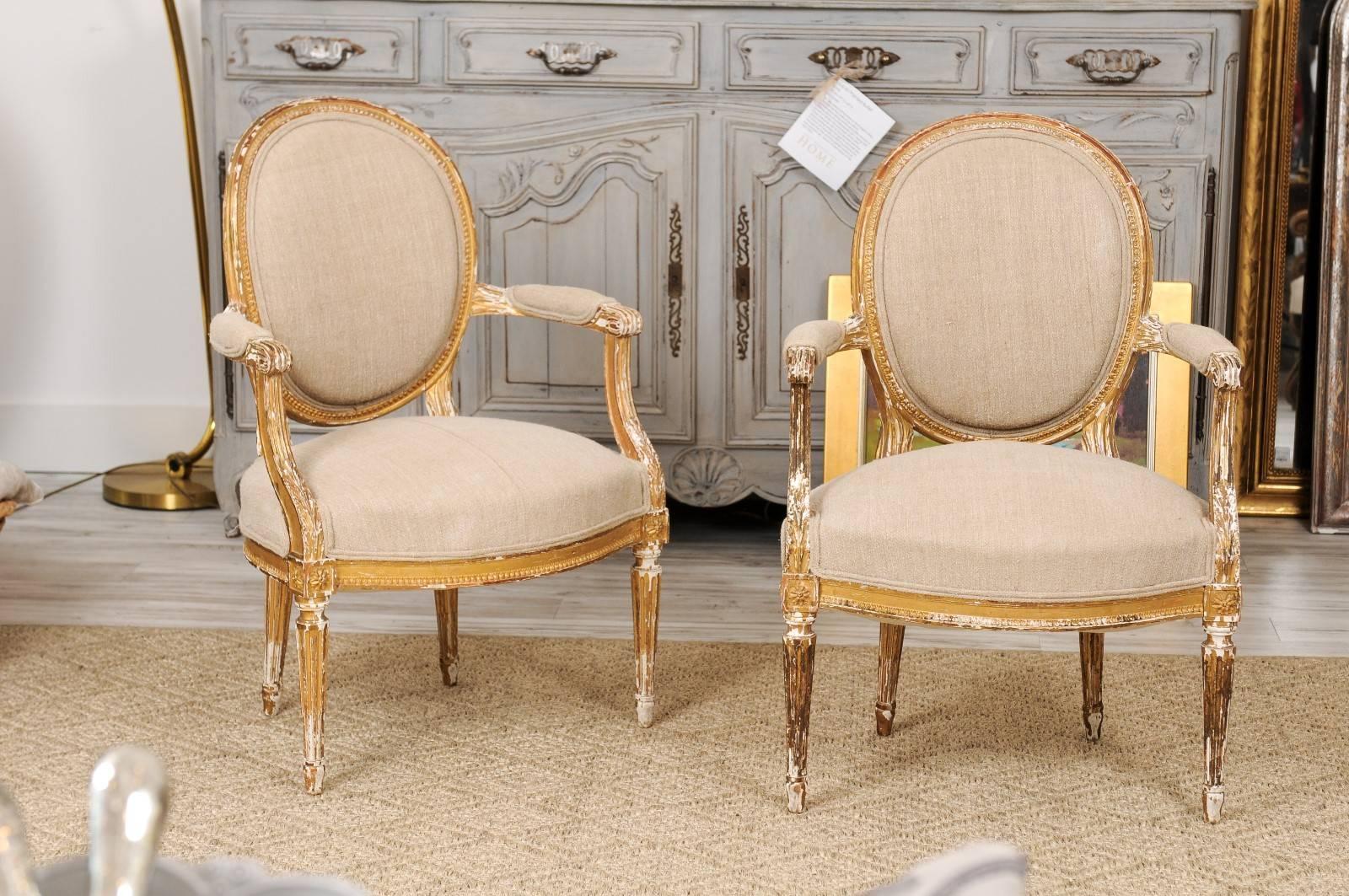 This pair of French Louis XVI style late 19th century giltwood armchairs feature upholstered oval backs with beaded moldings, partially upholstered scrolled arms, carved rosettes on the knees over four fluted and tapered legs. We often stay away